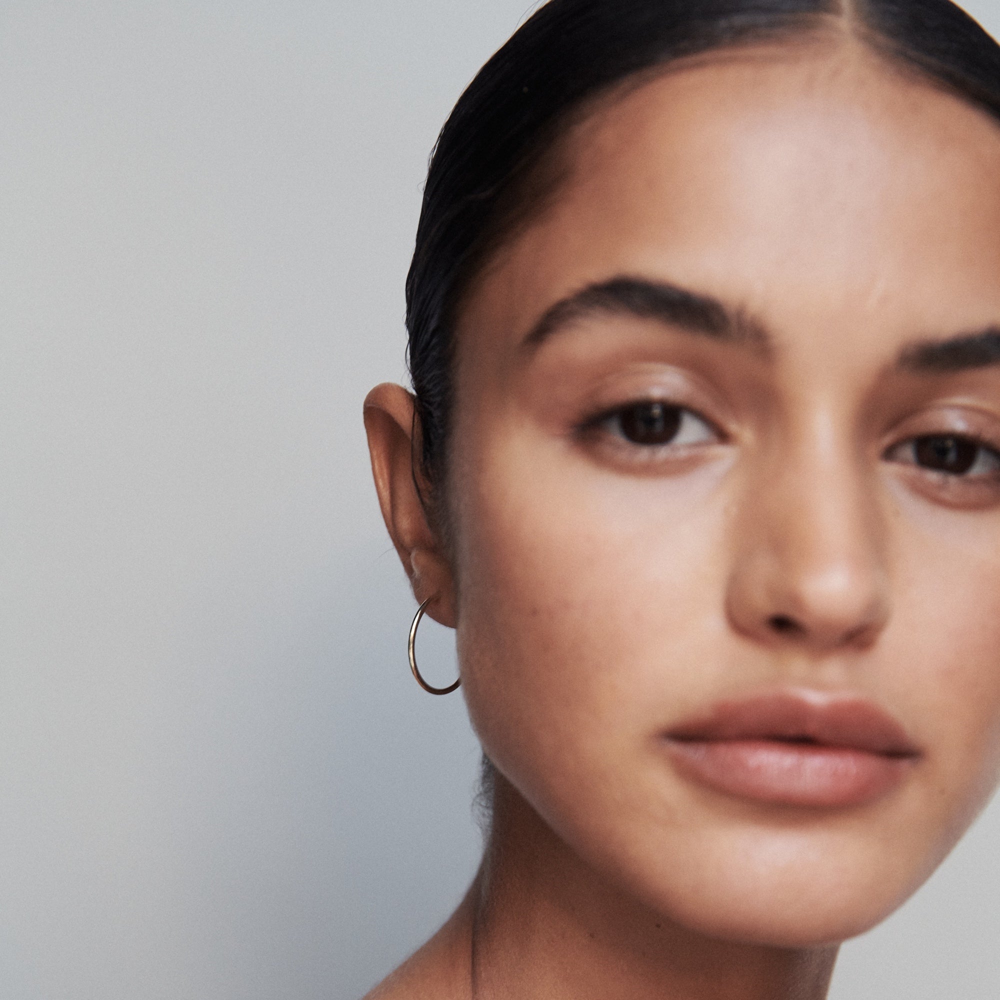 Woman wearing a sleek gold hoop earring, posing with a neutral expression against a plain background. Luxury jewellery focus. [text overlay: 15mm hoop] 
