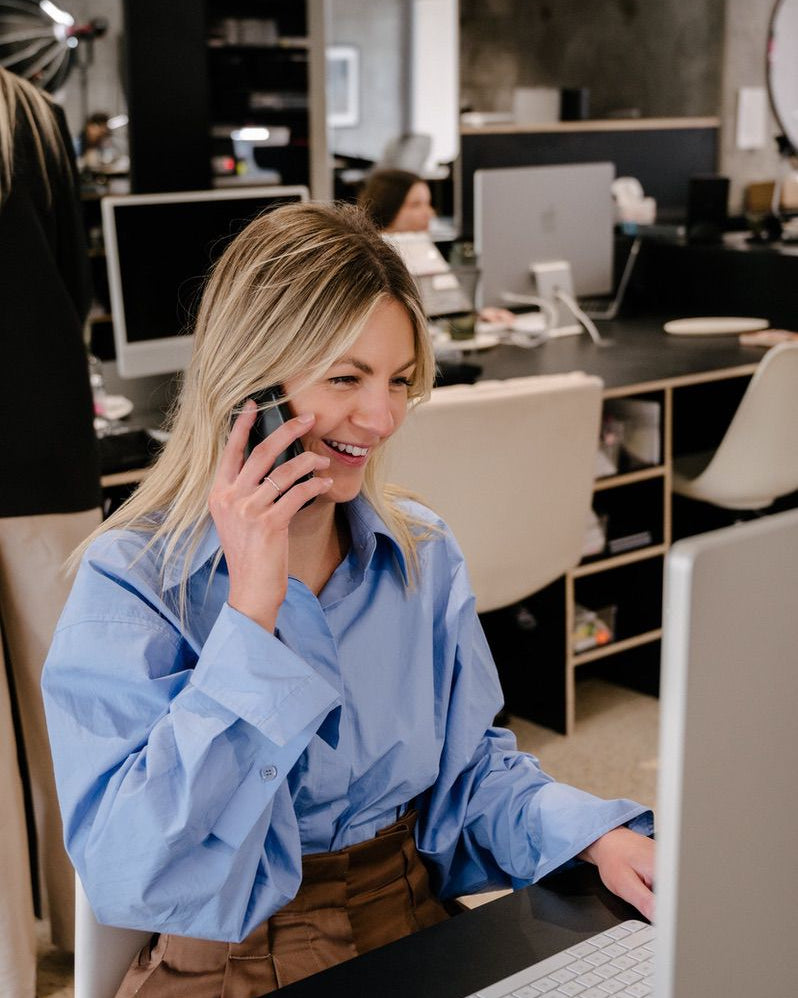 Woman in a blue shirt smiling while on the phone, working at a computer in a modern jewellery design office.