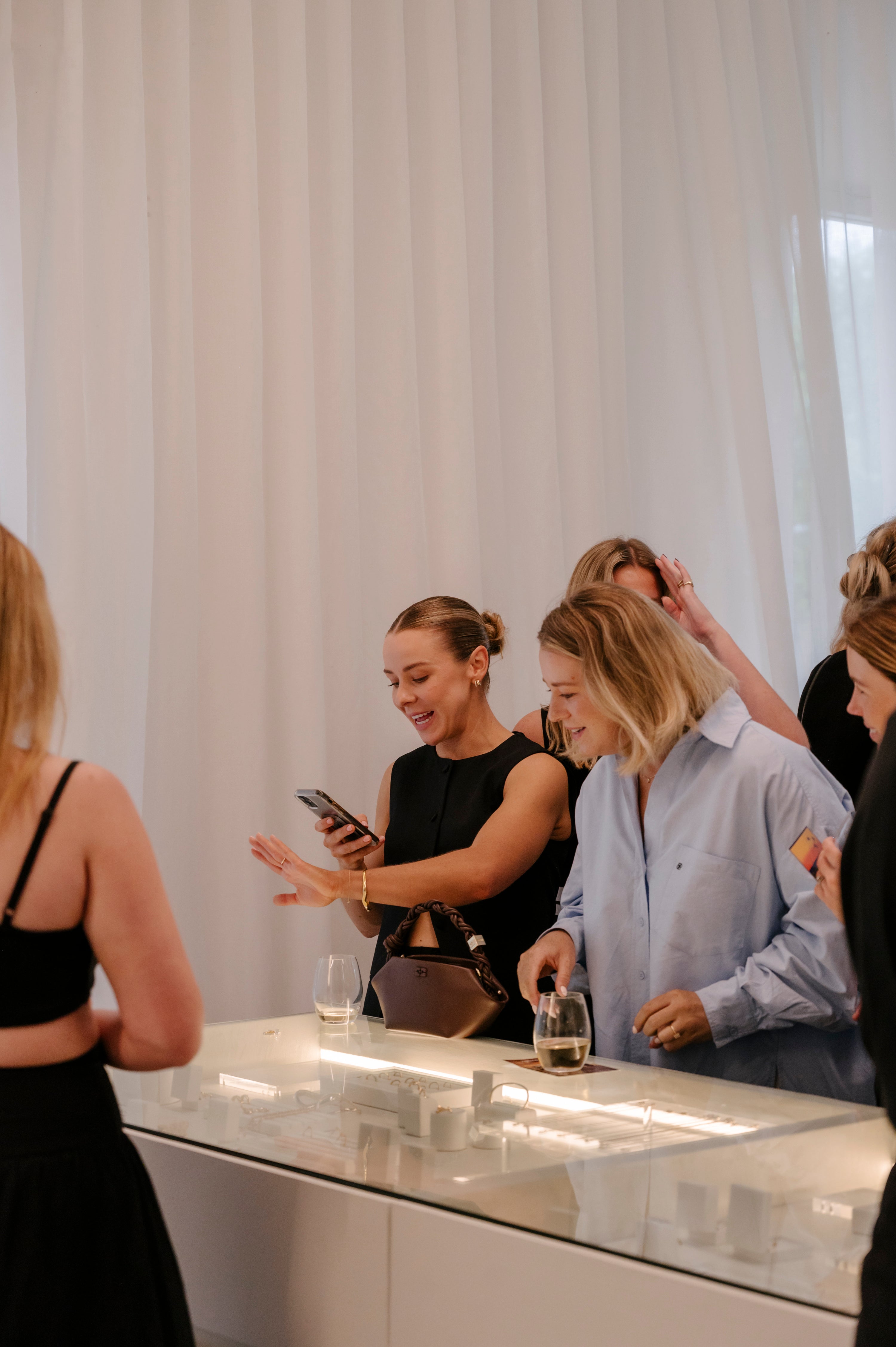 Women gathered around a luxury jewellery display, admiring pieces and chatting, with drinks in hand against a backdrop of elegant white curtains.