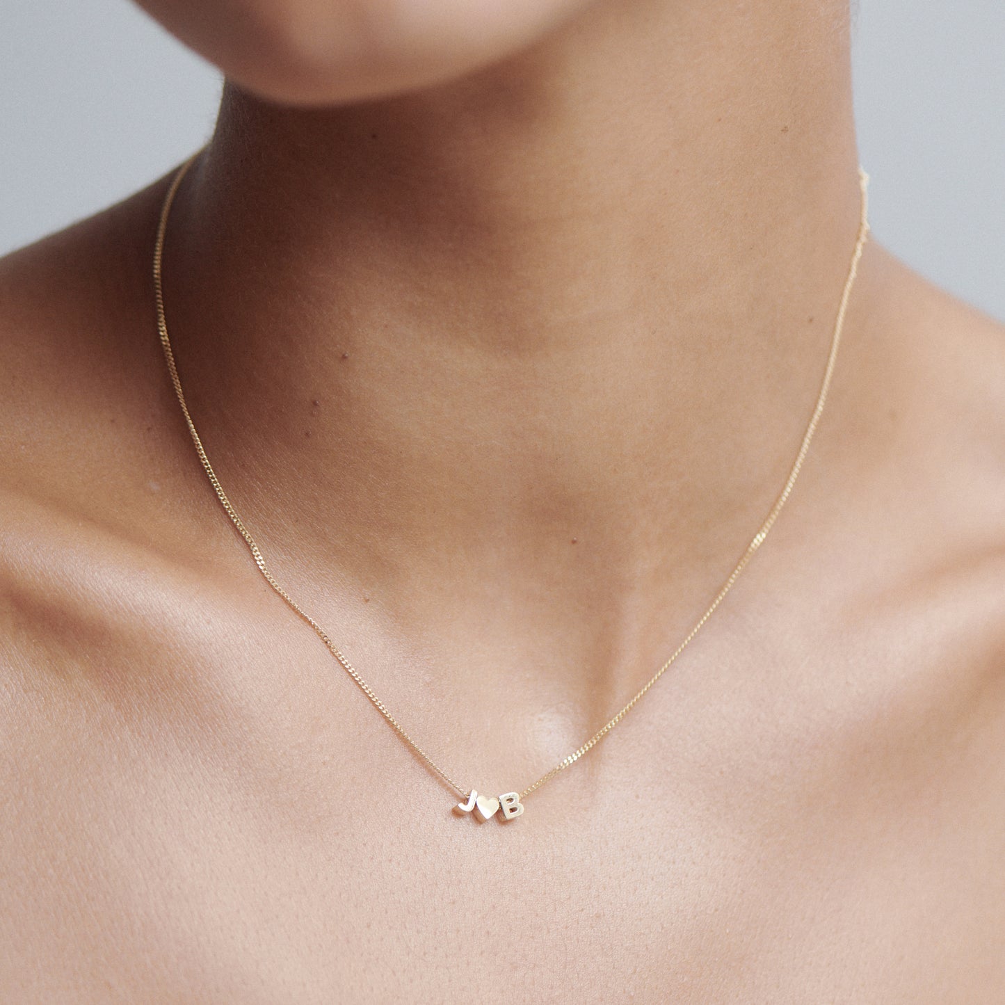 Close-up of a gold necklace with small heart and 'B' charms, worn on a person's neck. Minimalist luxury jewellery design.