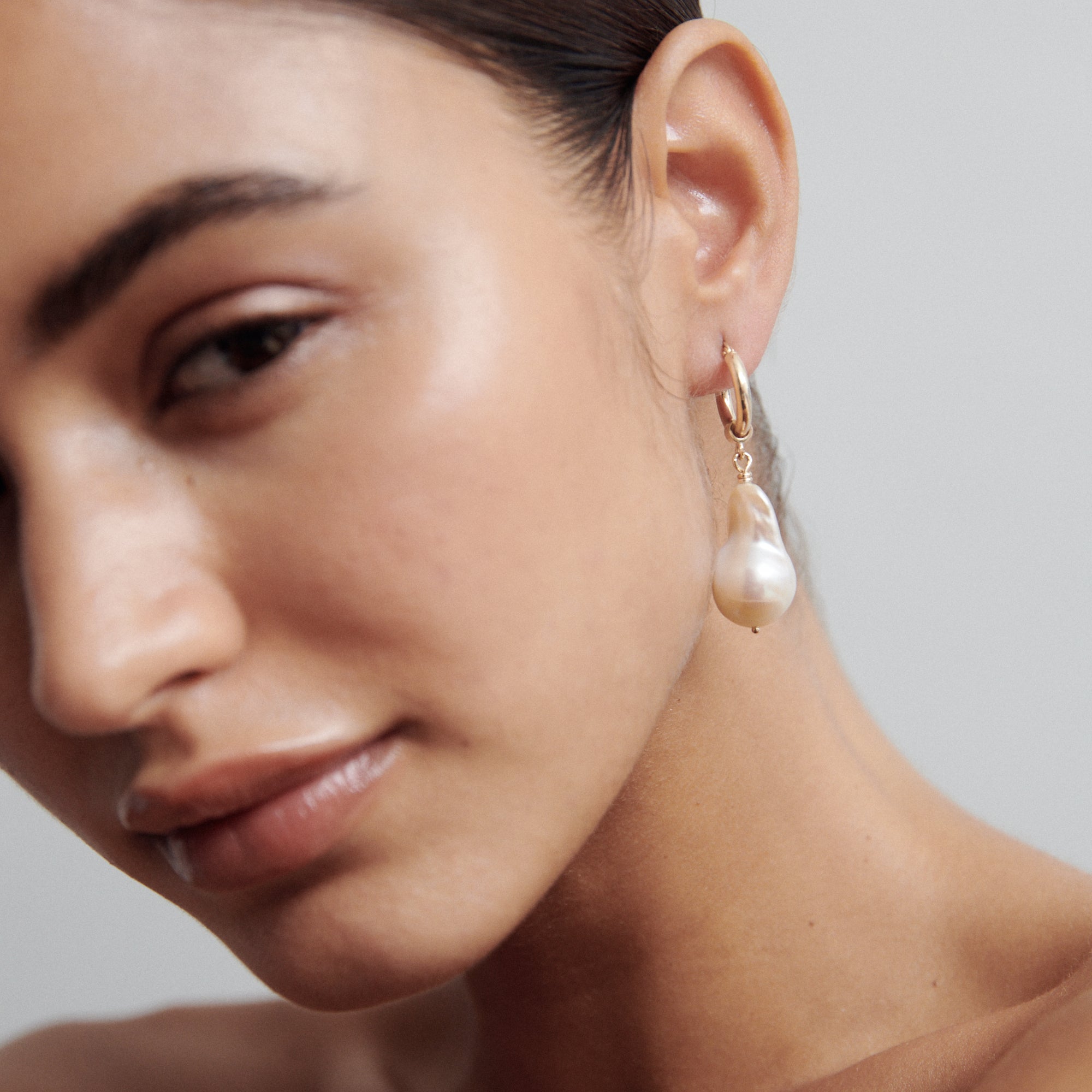 Close-up of a woman wearing a gold hoop earring with a dangling baroque pearl, showcasing luxury jewellery. Soft lighting enhances elegance.