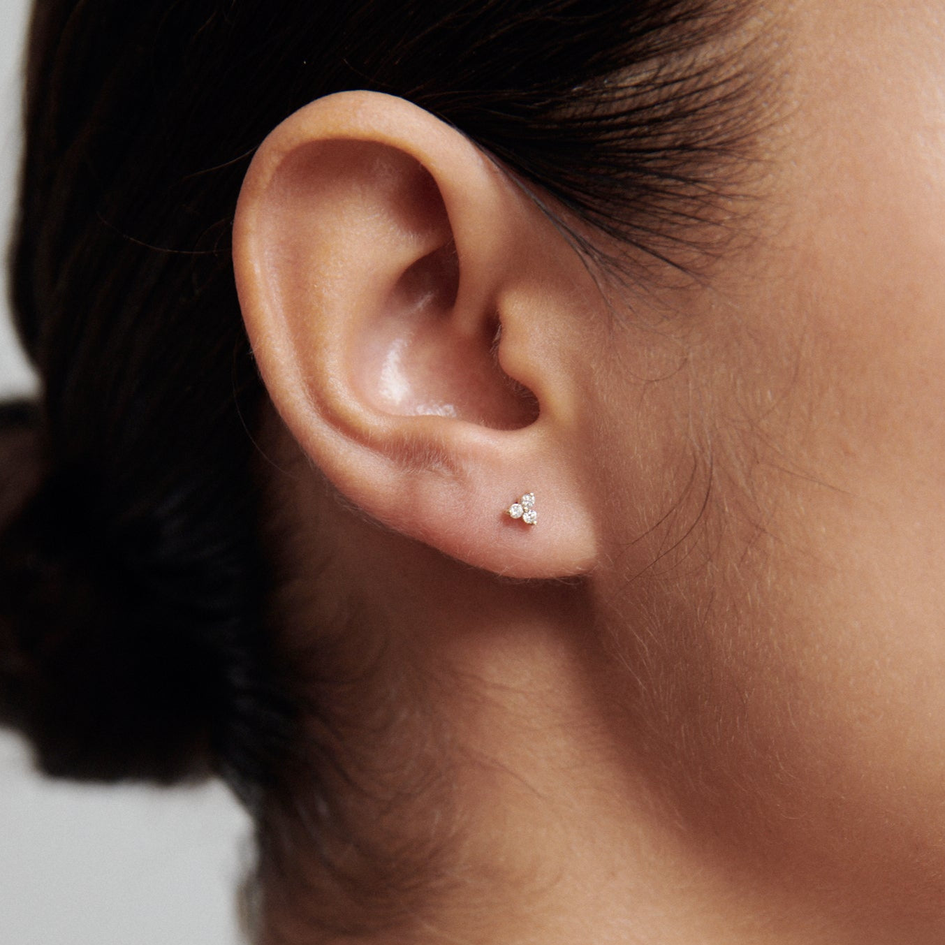 Woman's ear with a small diamond stud earring, hair styled in a neat bun, against a neutral background; luxury jewellery.