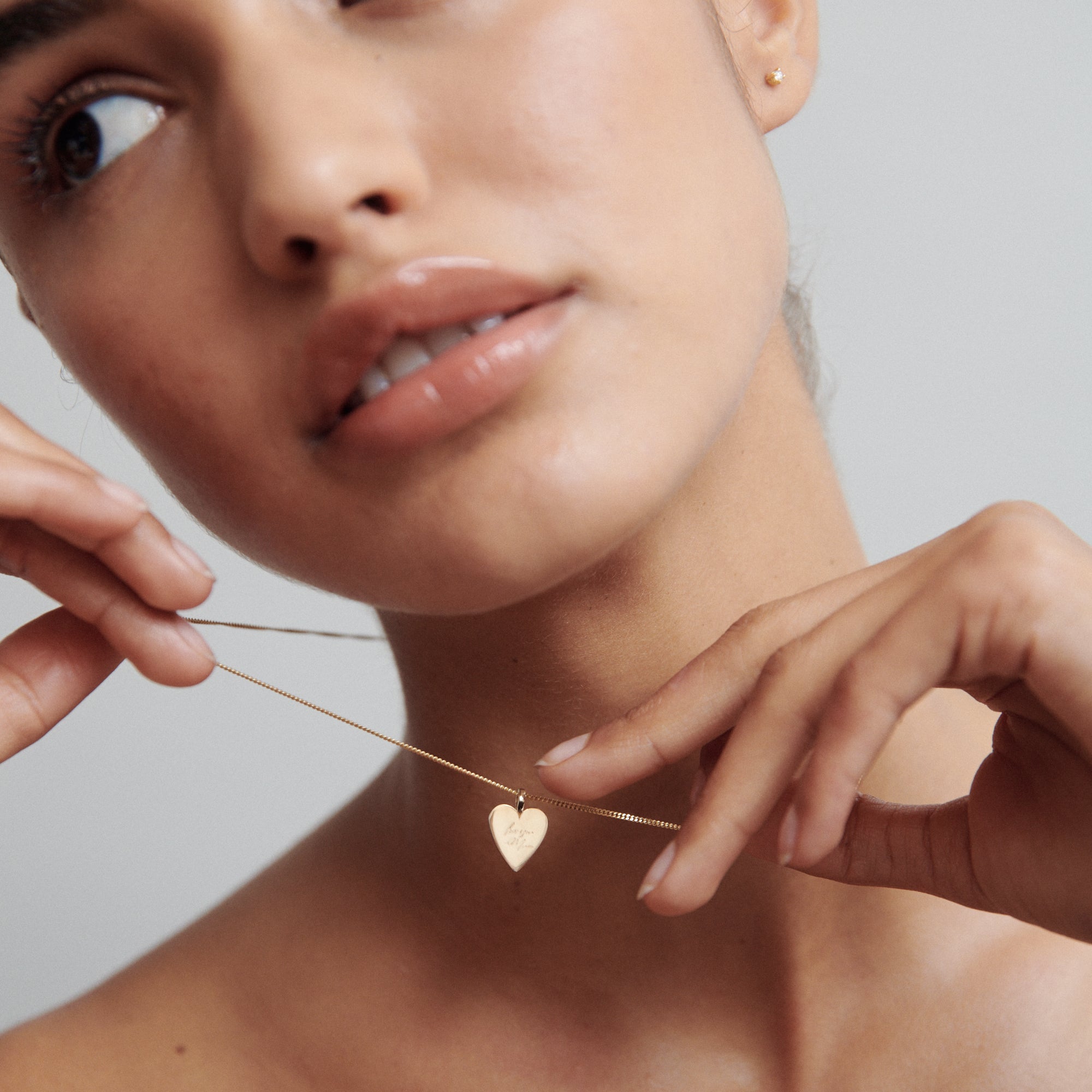 Close-up of person holding a gold heart necklace near their neck against a neutral background. Minimalist luxury jewellery style.[text overlay: ] 

