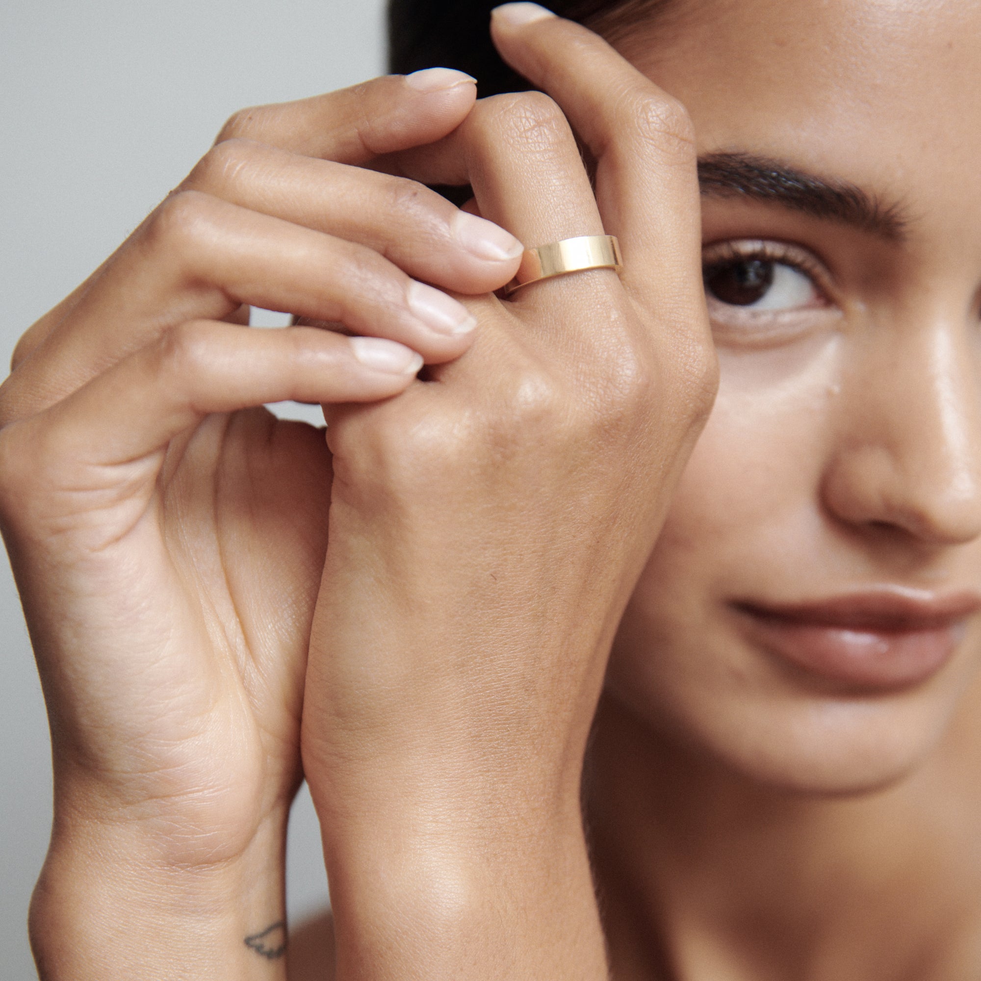Woman displaying an elegant gold ring on her finger, showcasing luxury jewellery. Her hand is gently touching her face, highlighting the ring's sleek design.  [text overlay:]
