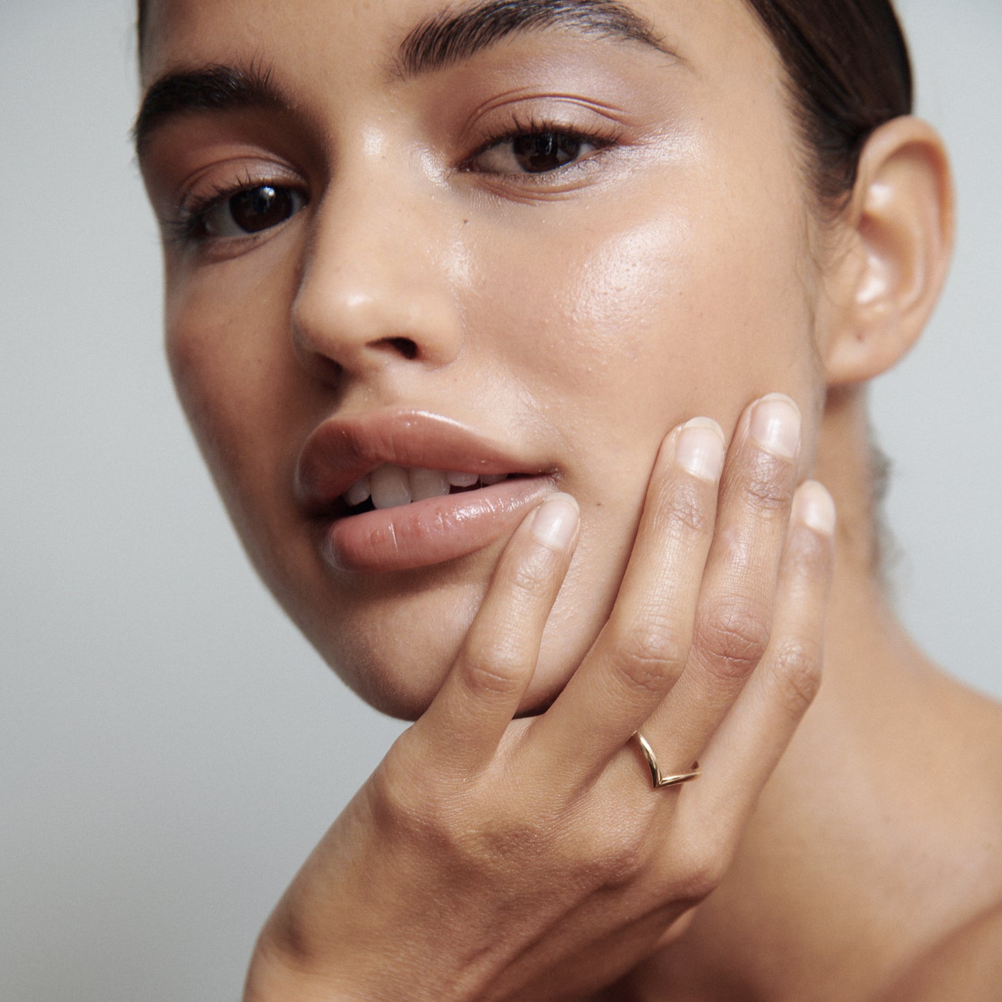 Woman with natural makeup, wearing a sleek gold ring, touches her face gently. Luxe jewellery, minimalist style.