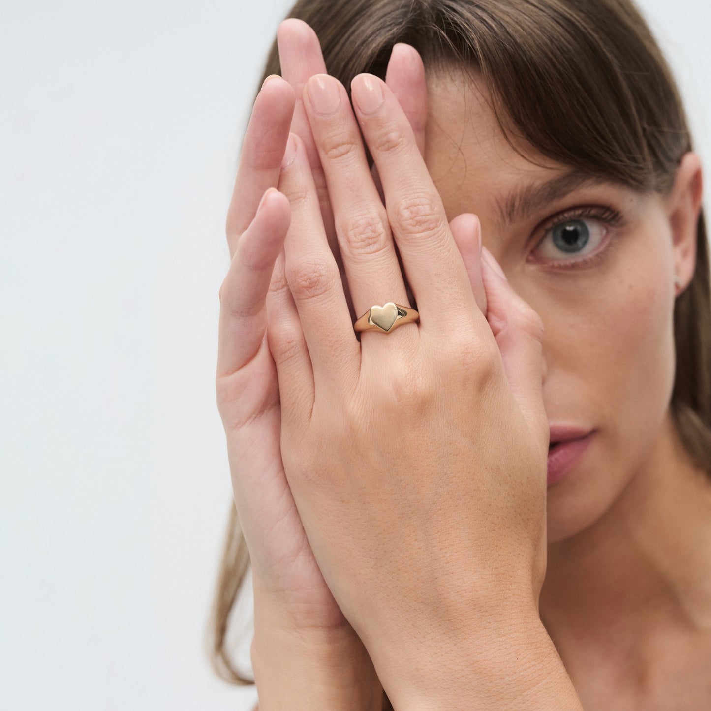 Woman with brown hair, holding hands up displaying a gold heart-shaped ring against her face, conveying elegance and luxury jewellery.