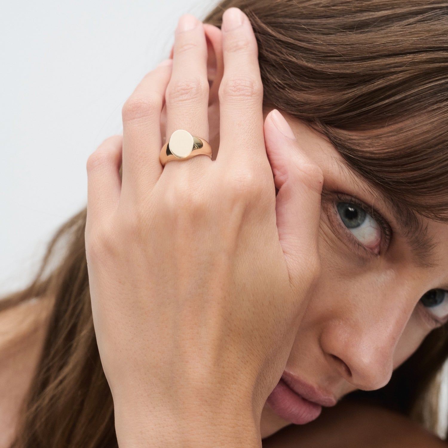 Woman wearing a sleek gold signet ring, close-up of her hand touching her face. Luxurious and elegant jewellery style.