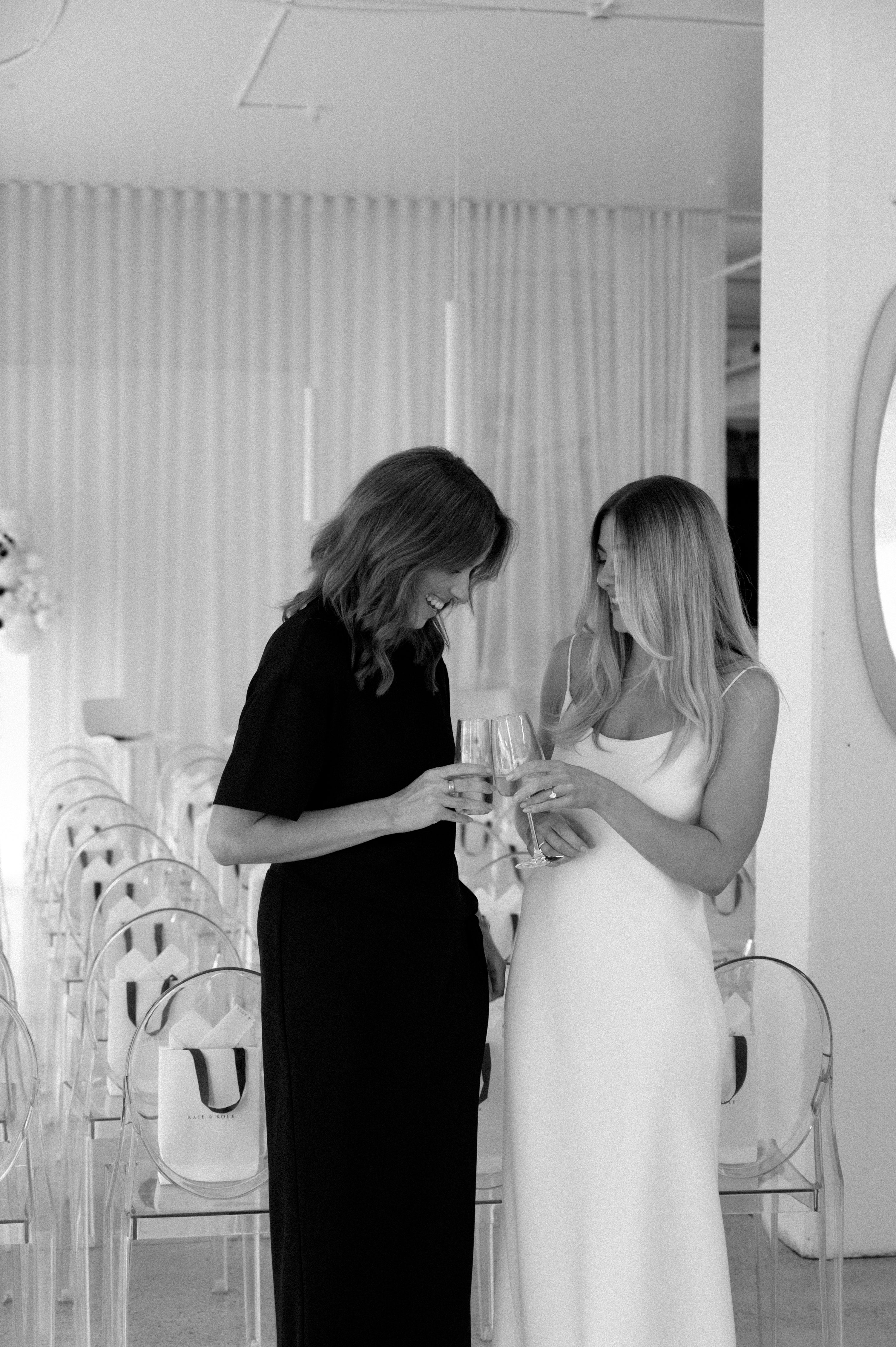 Two women in elegant dresses clink glasses in a chic, minimalist setting with clear chairs and draped curtains.