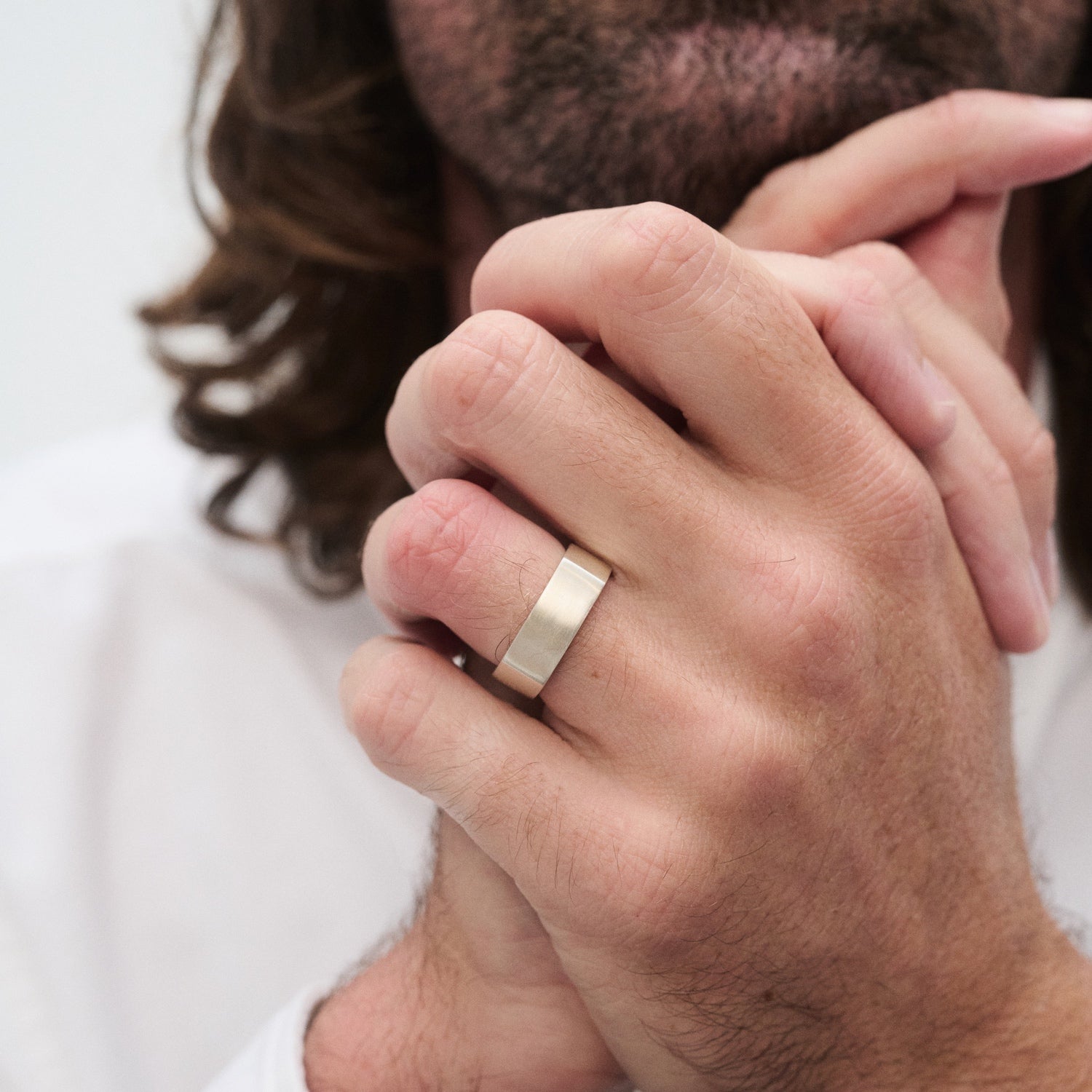 Man with long hair wearing a gold ring on his finger, hands clasped in front of a white shirt. Elegantly styled luxury jewellery focus.