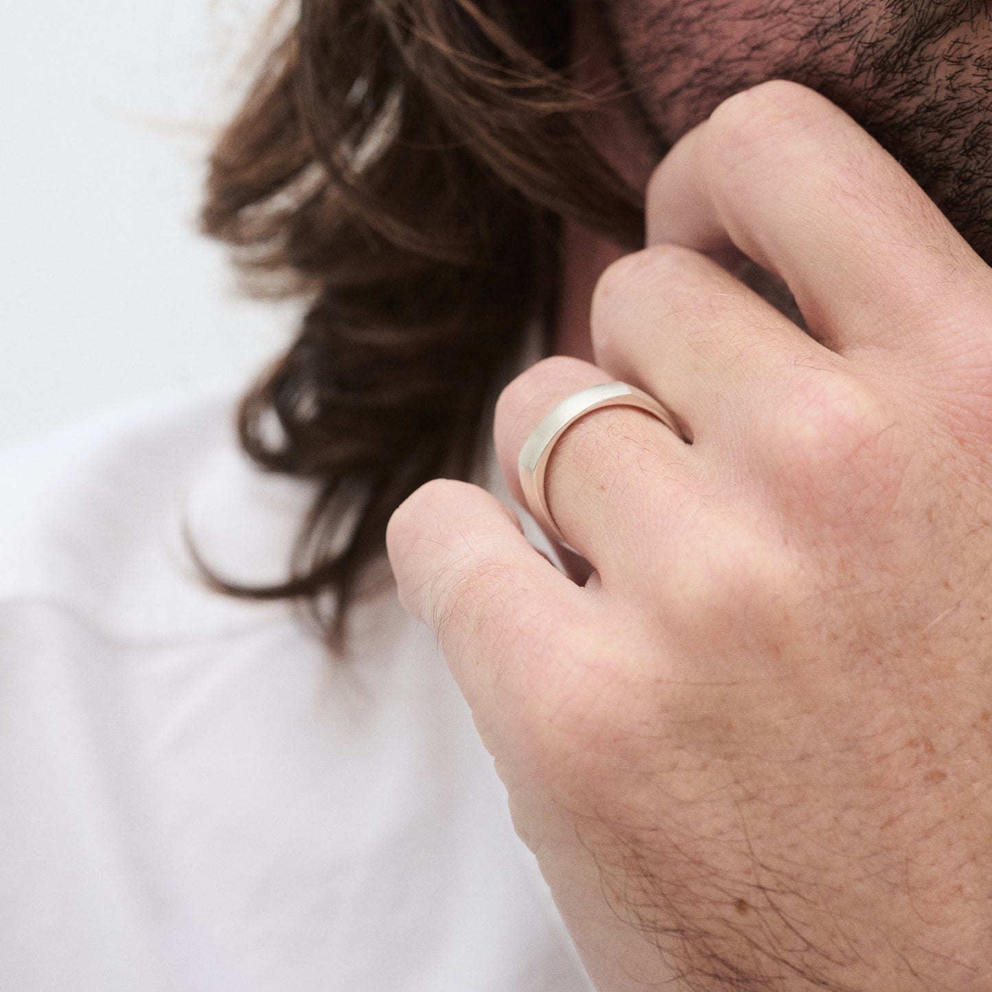 Man with long hair wearing a silver band ring on his finger, touching his chin thoughtfully. White shirt in background.