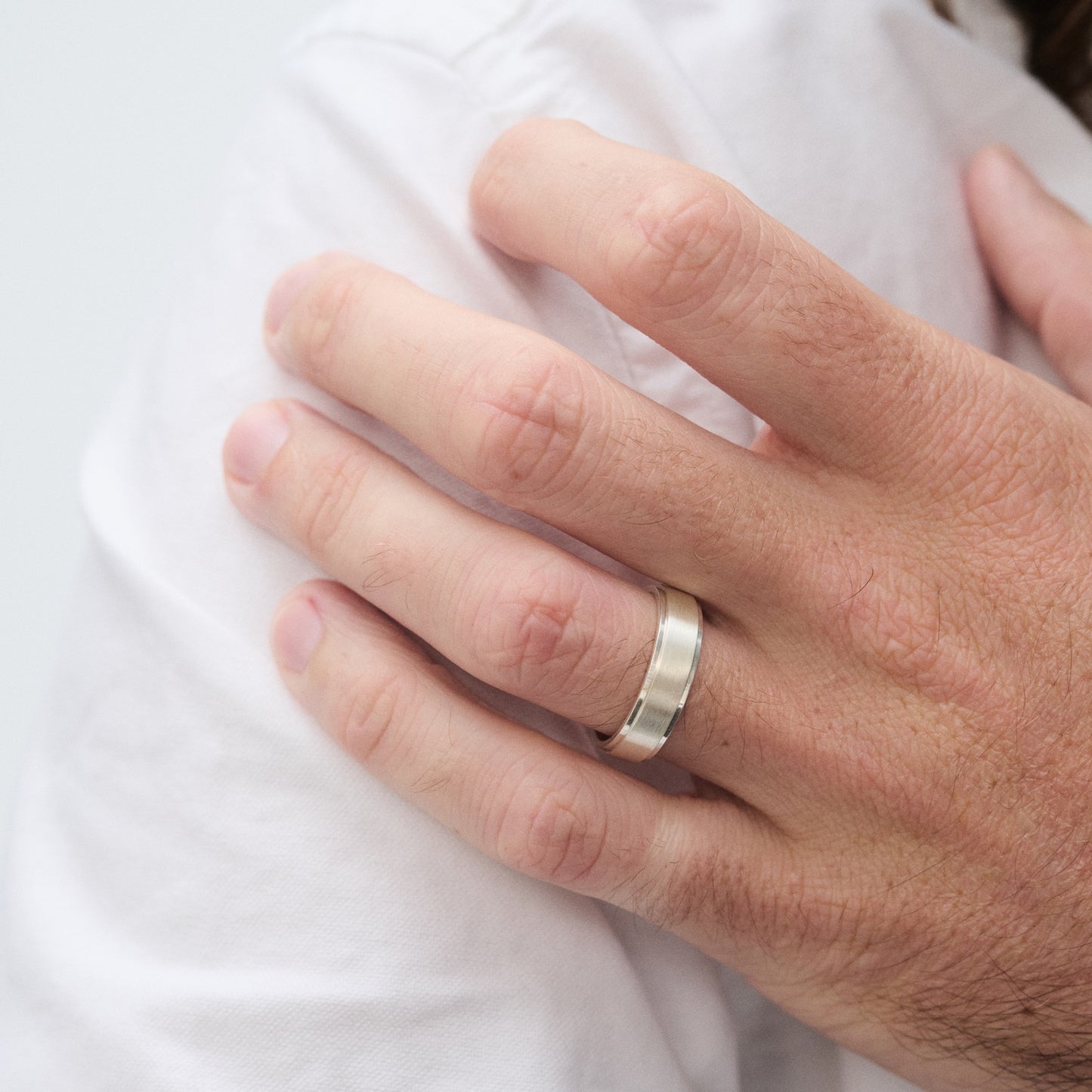Hand wearing an elegant silver band on ring finger, resting on a white shirt. Modern jewellery, luxury accessory, close-up.