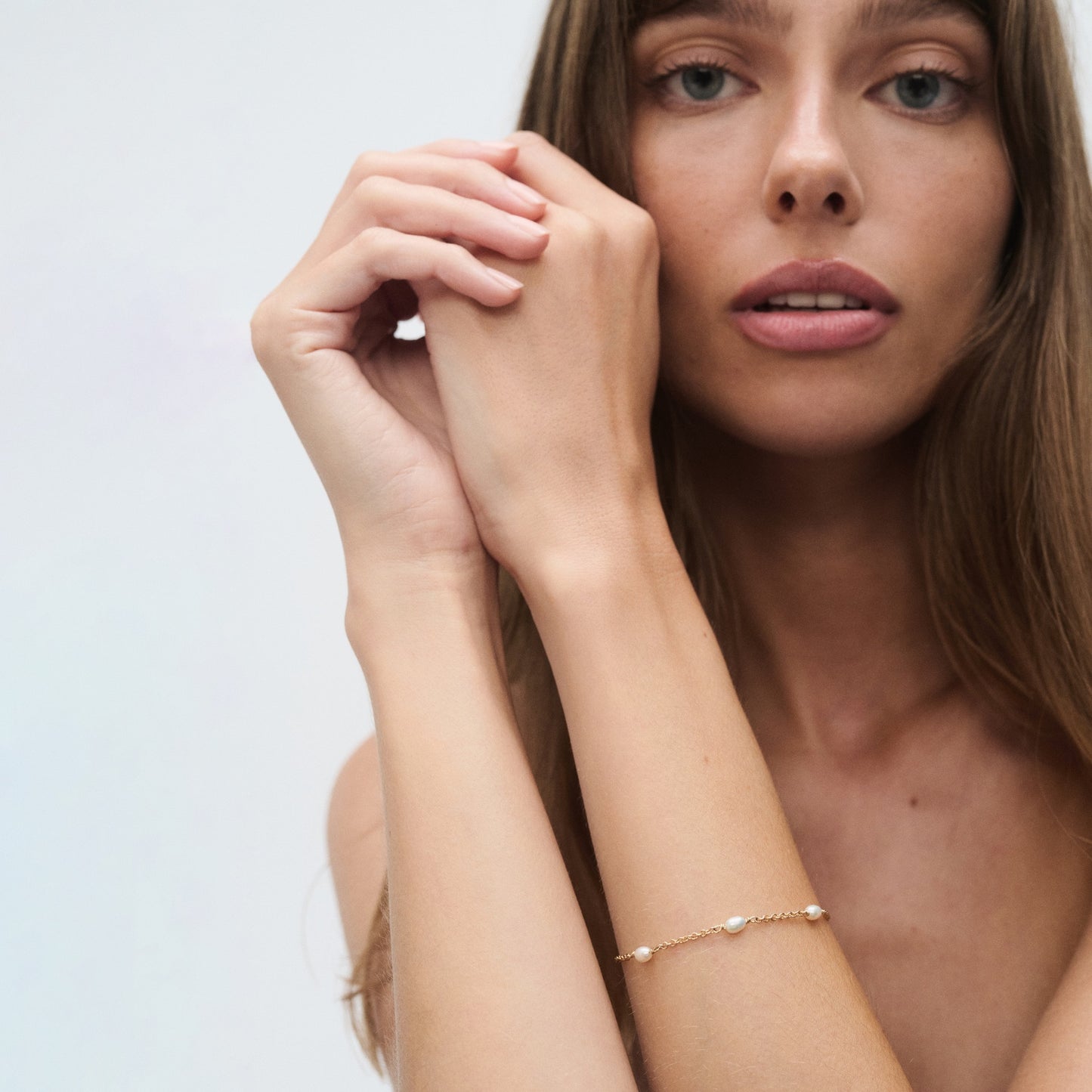 Woman with long brown hair wearing a delicate gold bracelet, posing with hands elegantly clasped near her face against a plain backdrop.
