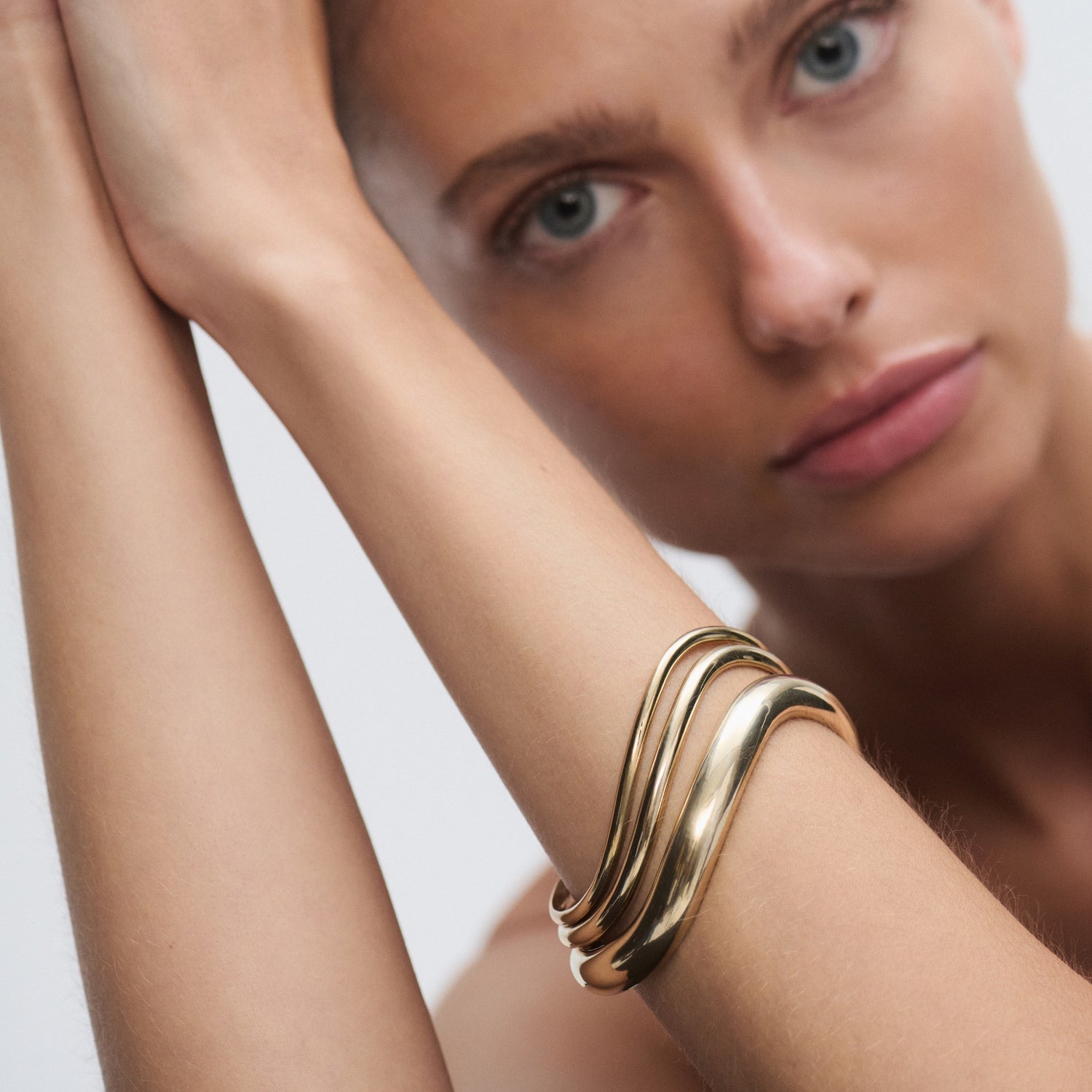 Model wearing an elegant, wavy gold bracelet; luxury jewellery close-up. Pale blue eyes, focused expression, soft background. [text overlay:] [text colour: #000000]
