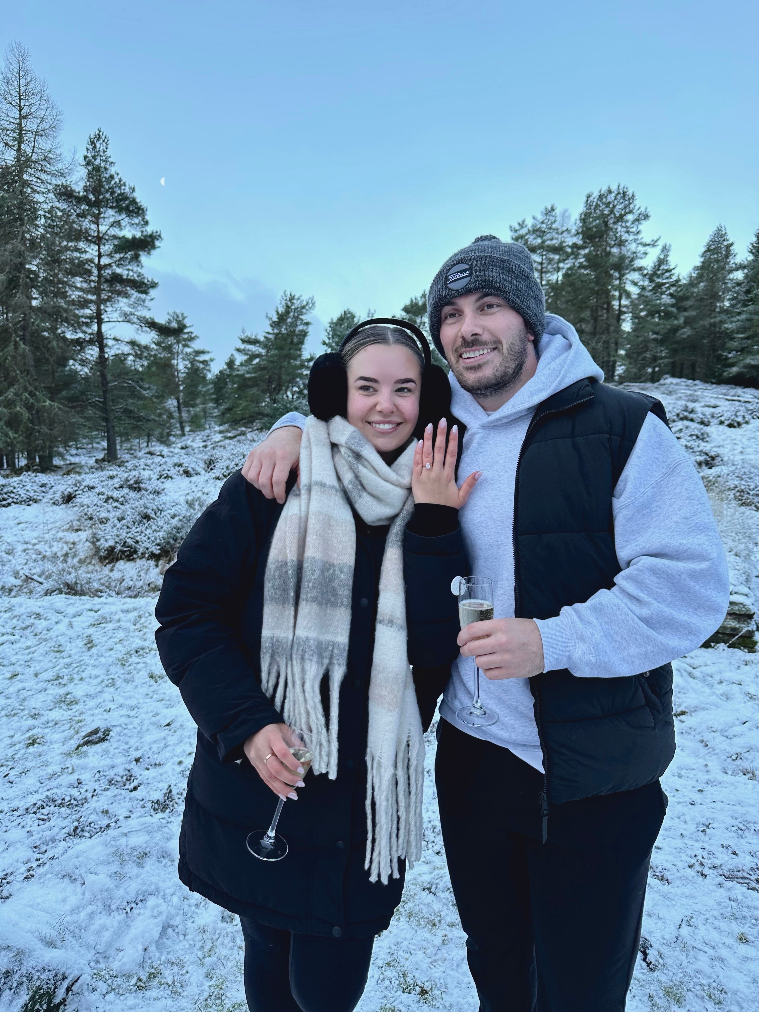 Couple celebrating engagement in snowy landscape, woman showing ring, both holding champagne flutes, wearing winter clothing.
