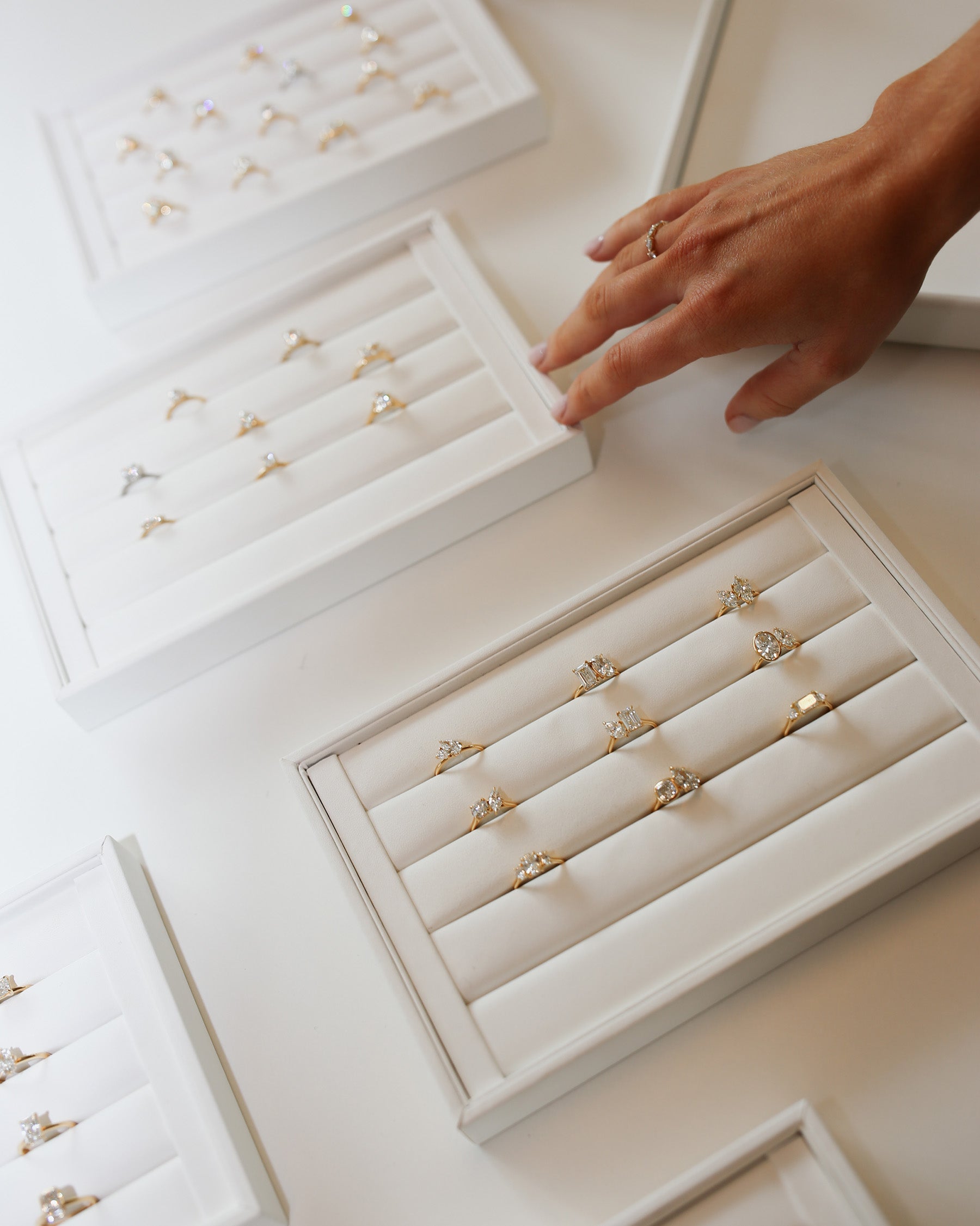 A hand reaching for gold and diamond rings elegantly displayed in white trays, showcasing high-end luxury jewellery.