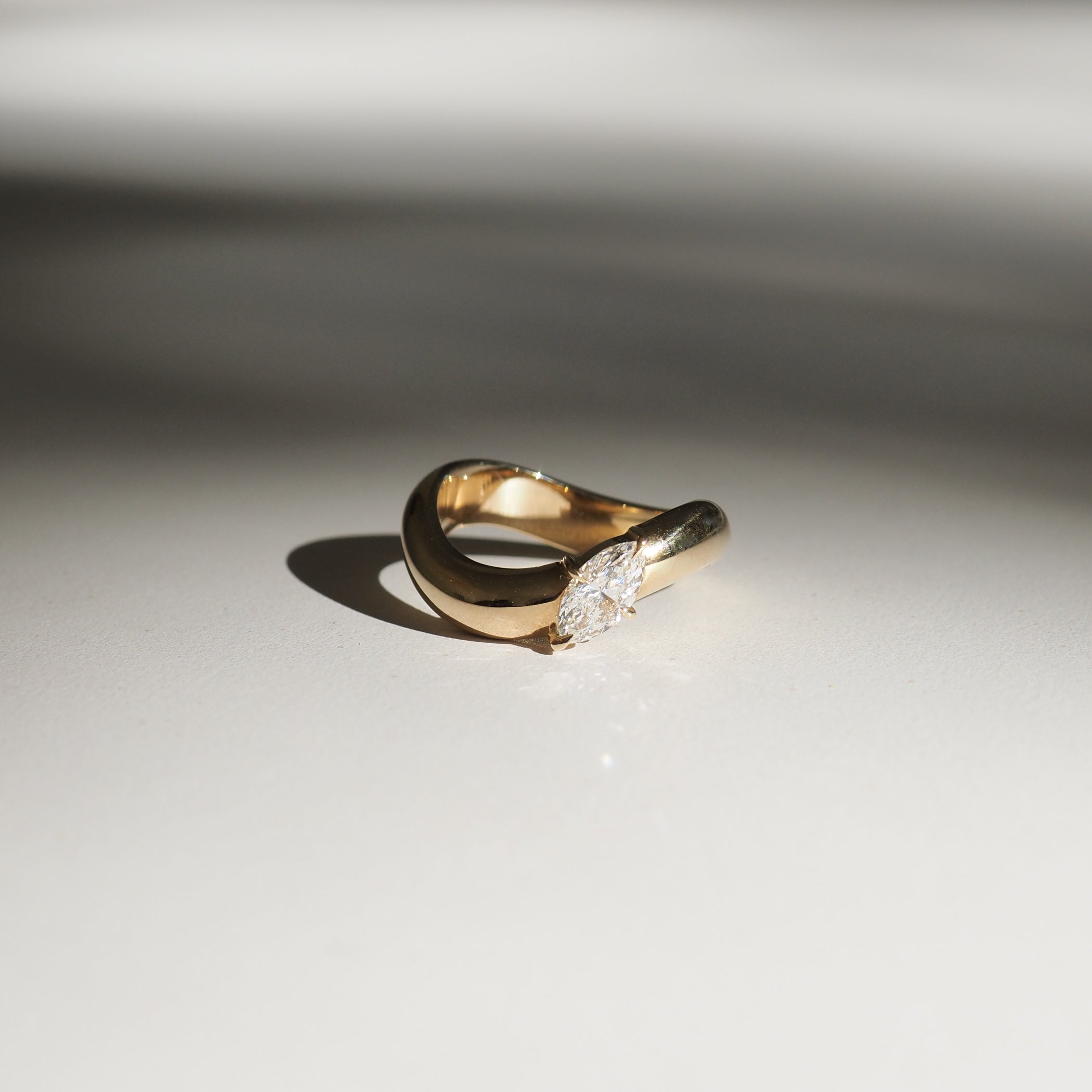 Gold ring with a marquise diamond set in a unique twist design, casting a soft shadow on a neutral surface. Luxurious and elegant jewellery.