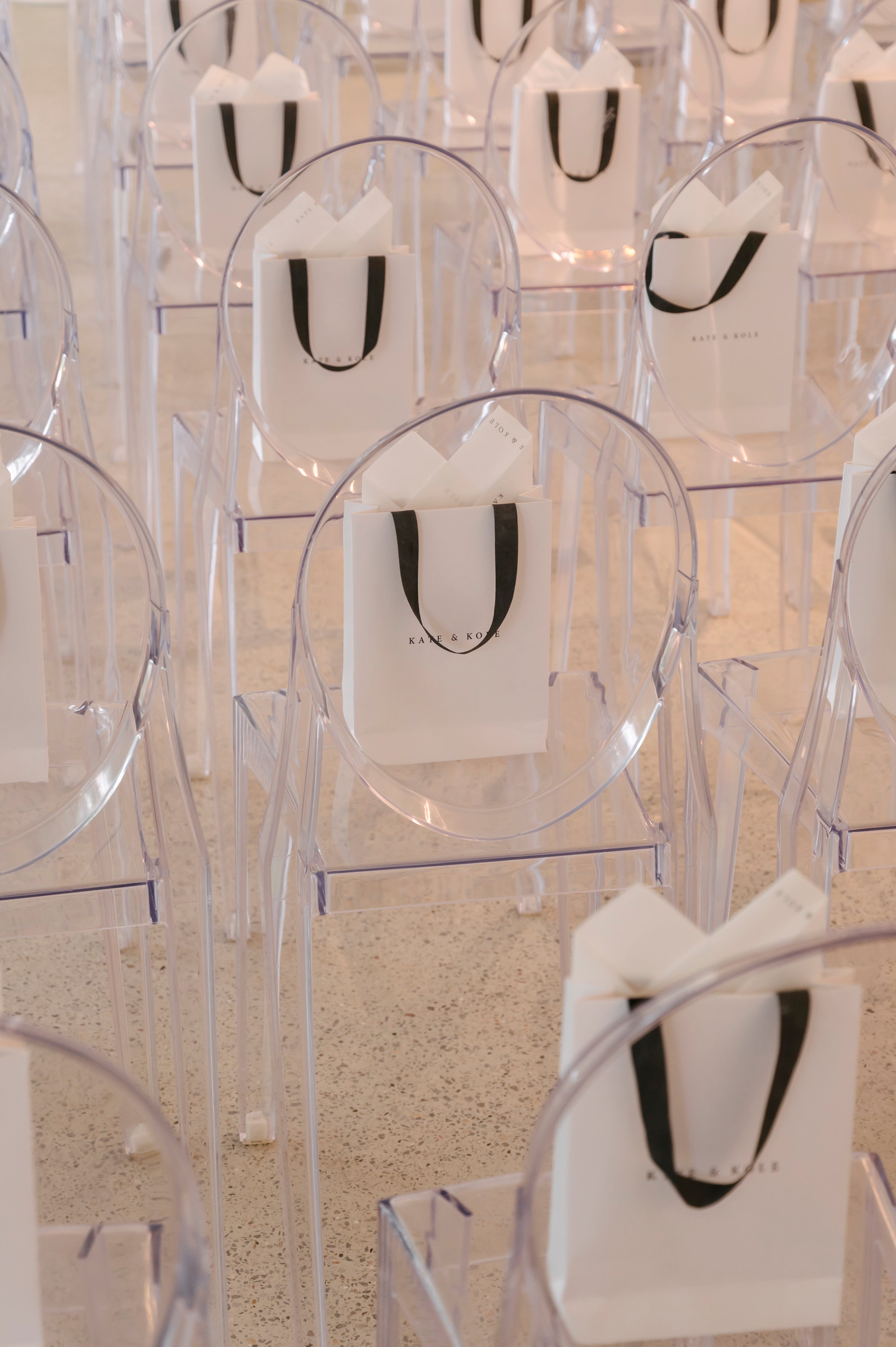 Rows of elegant clear chairs, each with a white luxury gift bag featuring black handles, set on a polished floor, suggesting a high-end event.