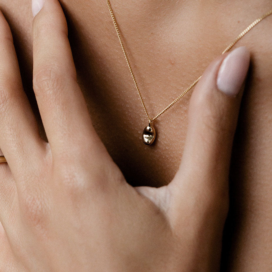Close-up of a hand touching a gold necklace with an elegant oval pendant, showcasing luxury jewellery detail and craftsmanship.