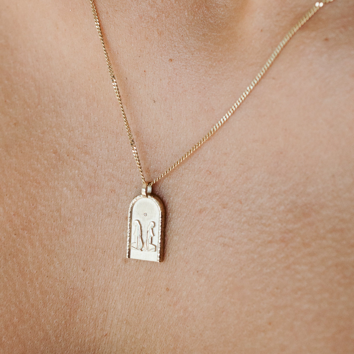 Gold necklace with an engraved pendant depicting abstract figures, worn on bare skin. Luxurious jewellery close-up detail.