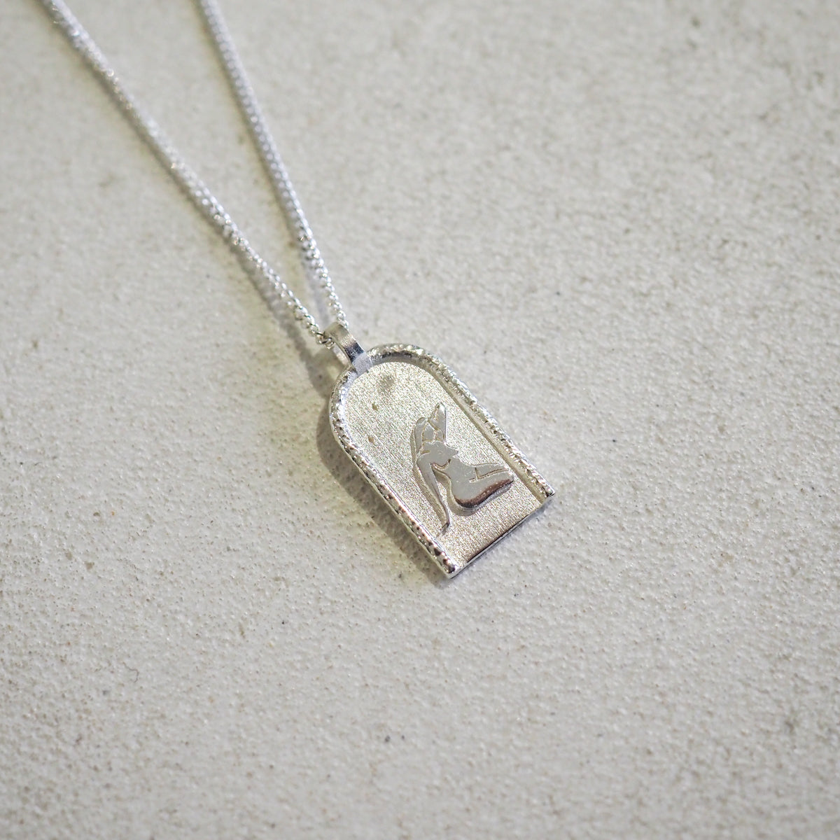 Silver pendant with an engraved angel motif on a fine chain, displayed on a light, textured surface. Elegant, luxury jewellery design.