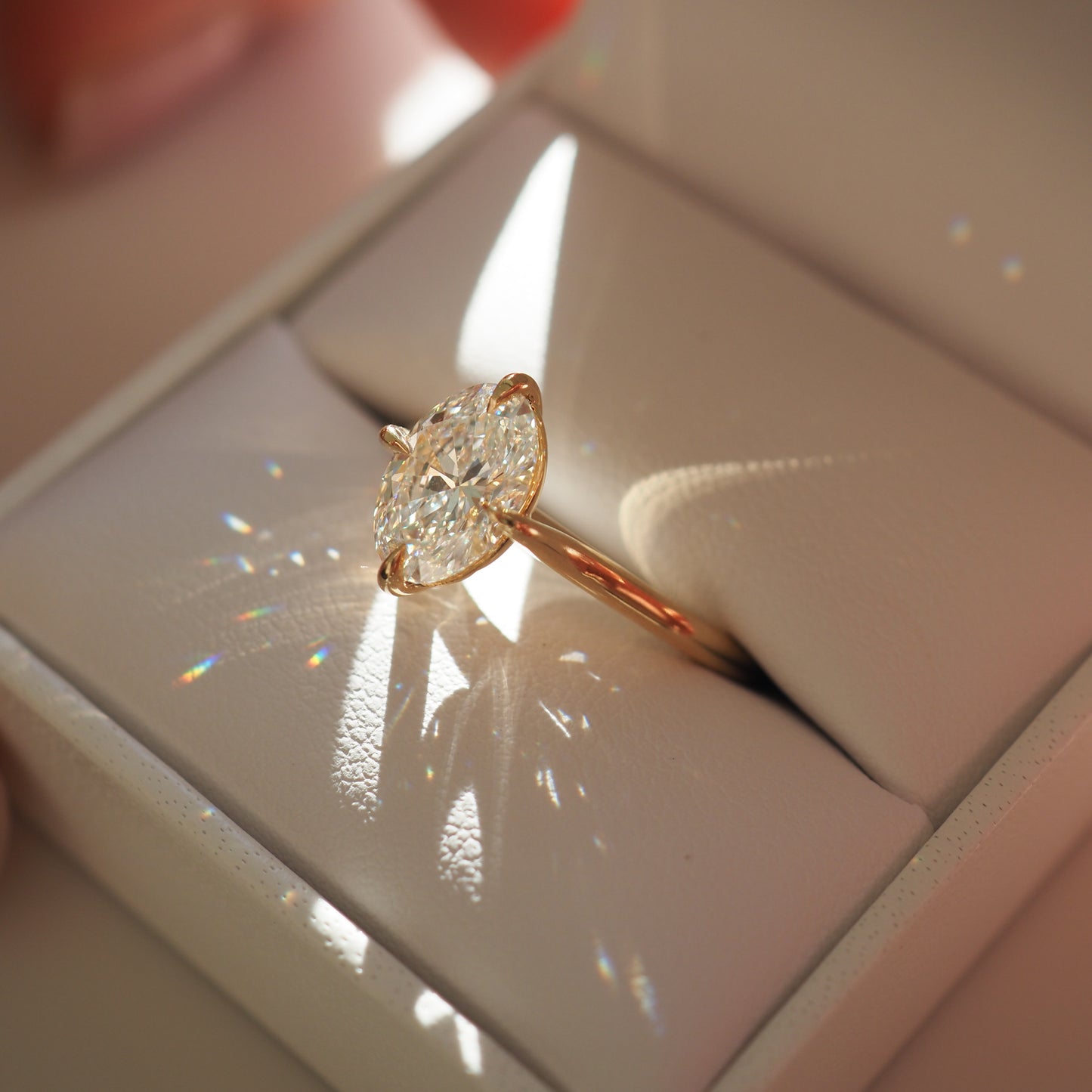 A marquise-cut diamond ring in a rose gold setting, placed inside a white jewellery box. Sunlight creates prism reflections around the ring.