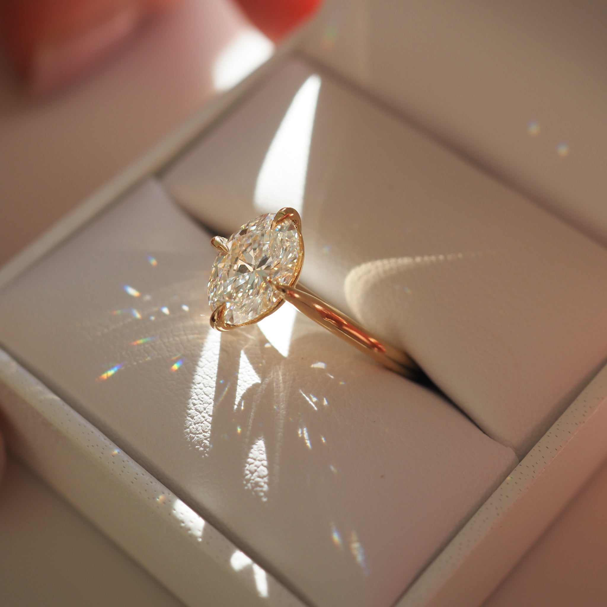 A marquise-cut diamond ring in a rose gold setting, placed inside a white jewellery box. Sunlight creates prism reflections around the ring.