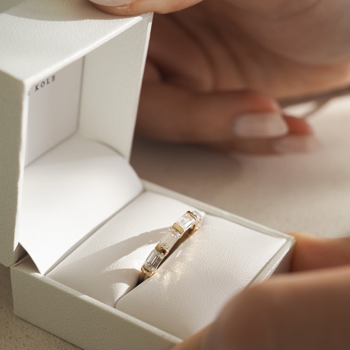Gold ring with square diamonds in a white jewellery box, held by manicured hands. Elegant and luxurious design.