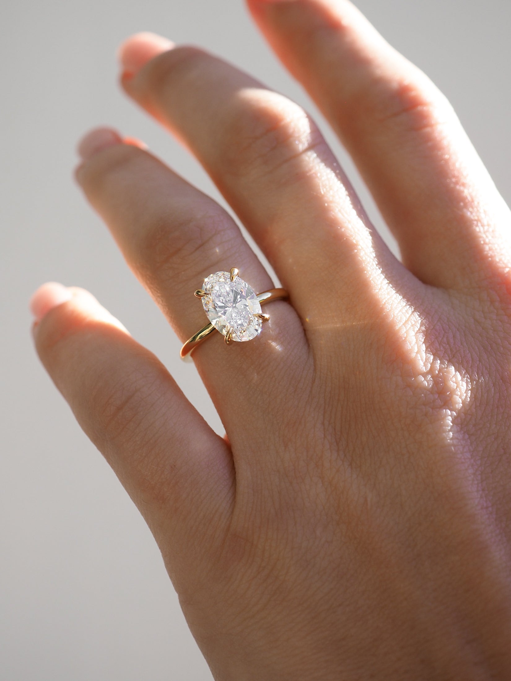 Hand adorned with an oval-cut diamond engagement ring on a gold band, reflecting sunlight. Luxury jewellery, high-end design.