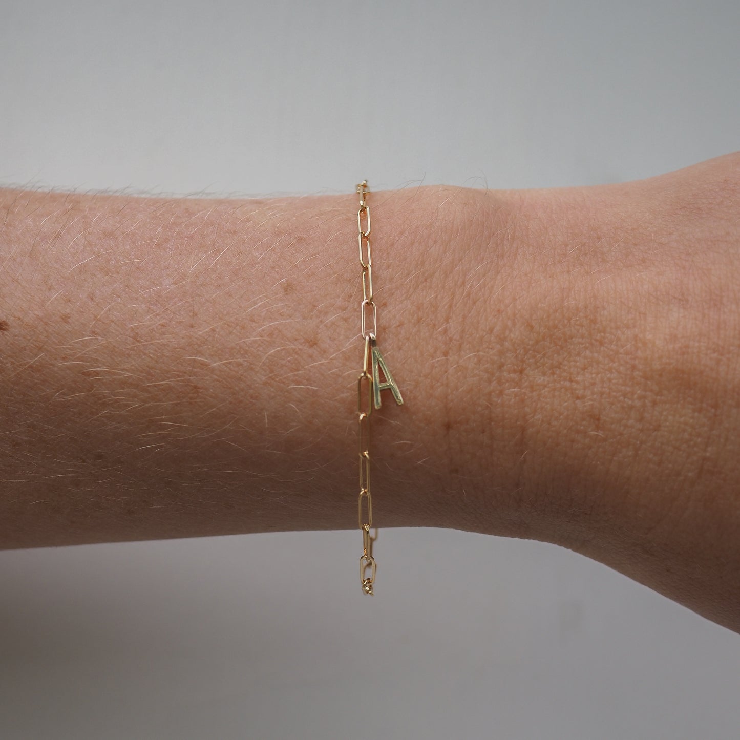 Gold chain bracelet with an 'A' initial pendant on a wrist, against a plain background. Minimalist, luxury jewellery design.