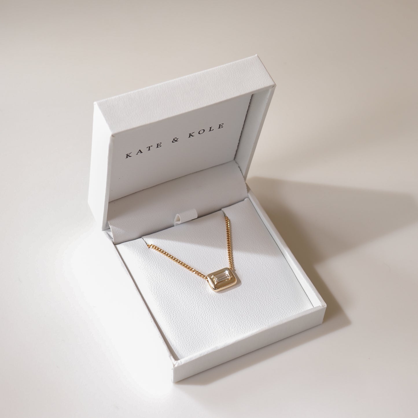 Gold necklace with a rectangular pendant in a white Kate & Kole jewellery box. Elegant, minimal design on a subtle cream background.