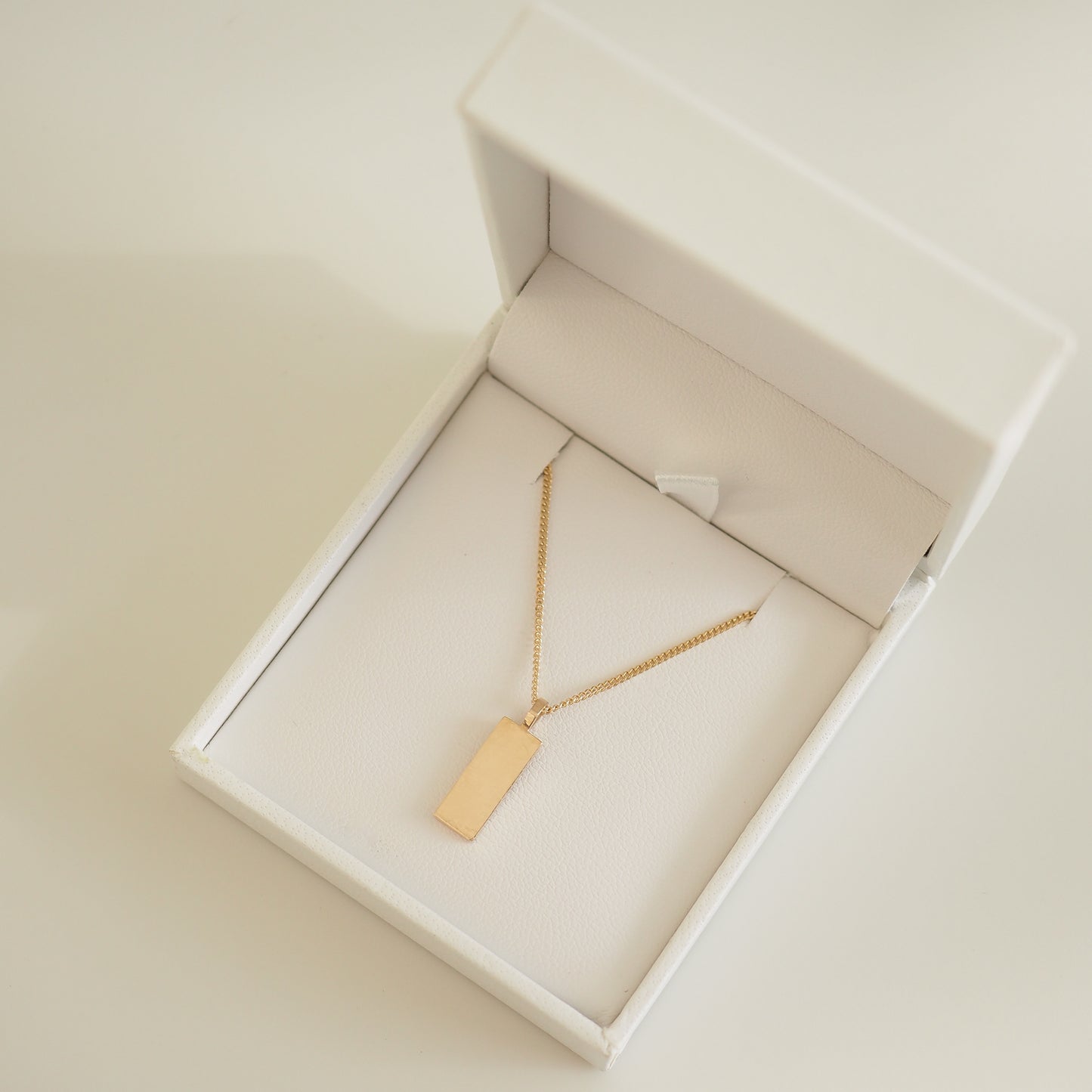 Gold rectangular pendant necklace in an elegant white jewellery box. Simple, luxurious design perfect for high-end fashion enthusiasts.