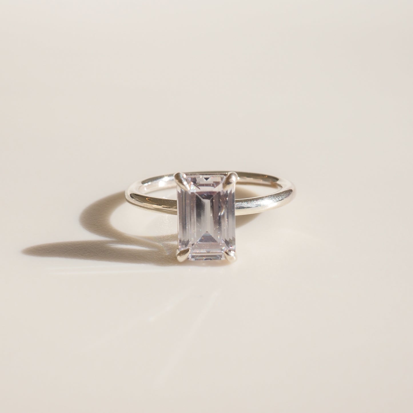 Elegant silver ring with a rectangular-cut clear gemstone, casting a shadow on a light surface; luxury jewellery photography.