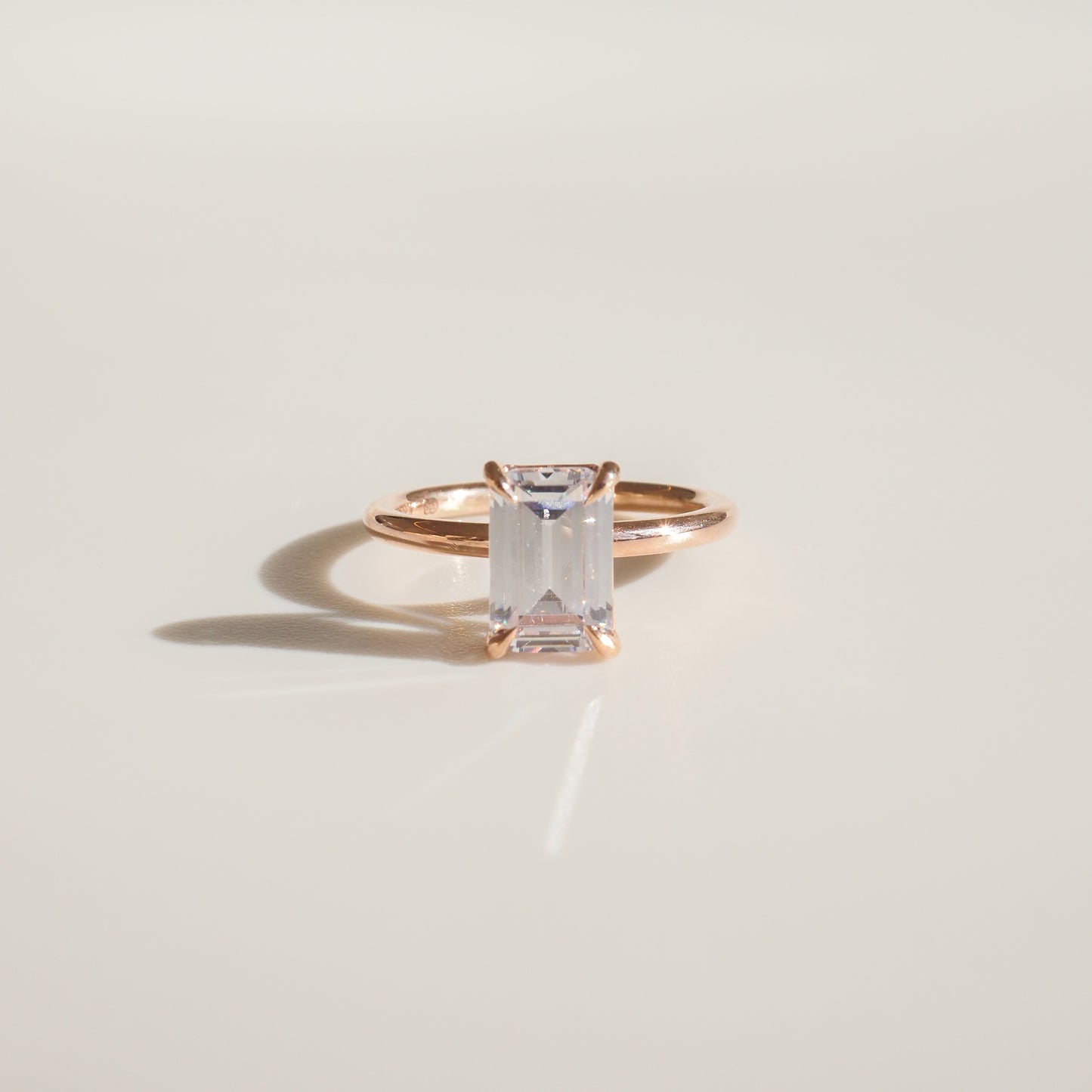 Emerald-cut diamond ring in rose gold setting on a soft beige background, casting a subtle shadow. Luxurious minimalist jewellery design.