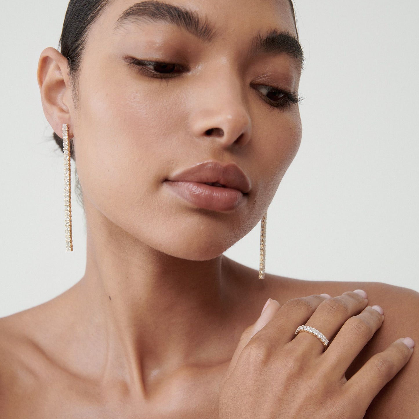 Woman wearing elegant gold drop earrings and a gold band ring adorned with small diamonds, against a neutral background.