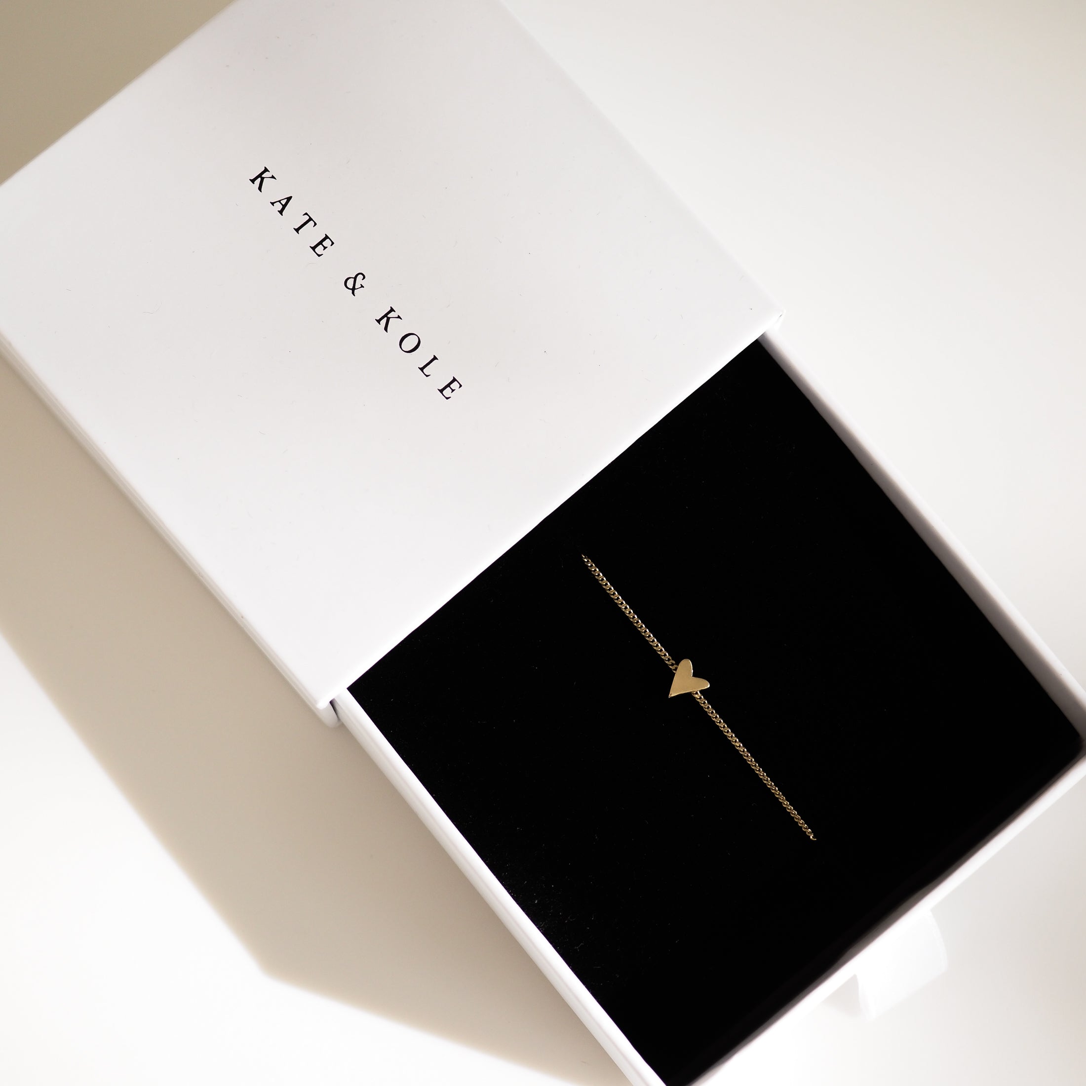 Gold heart bracelet in a Kate & Kole jewellery box, resting on black velvet fabric. Minimalist, luxury design.