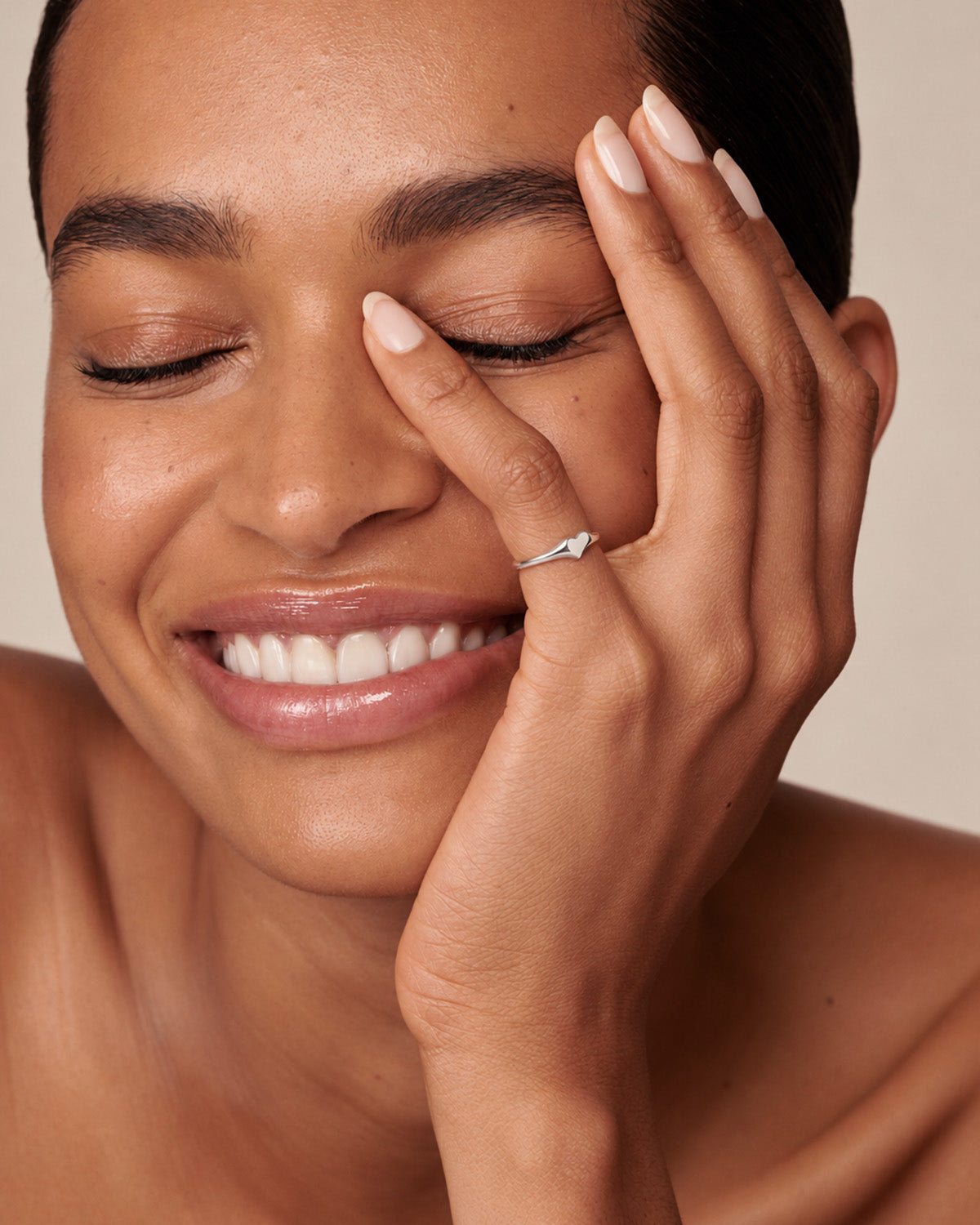 A close-up lifestyle shot of a woman with her eyes closed, smiling warmly while wearing a delicate silver ring on her finger.