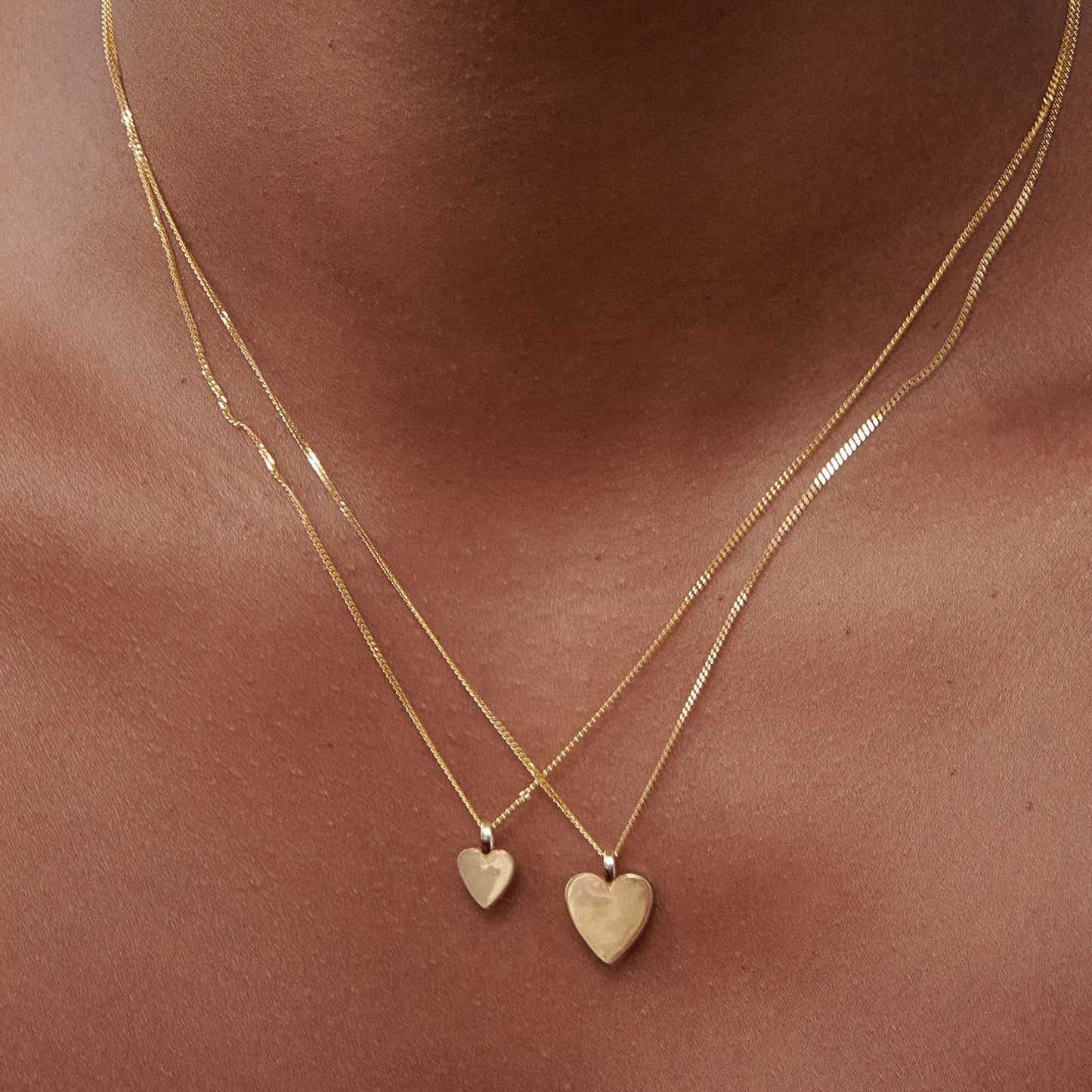 Close-up of a woman wearing two gold heart pendants on delicate chains against her skin, showcasing luxury jewellery design.[text overlay: Small pendant (left) and large pendant (right)] 