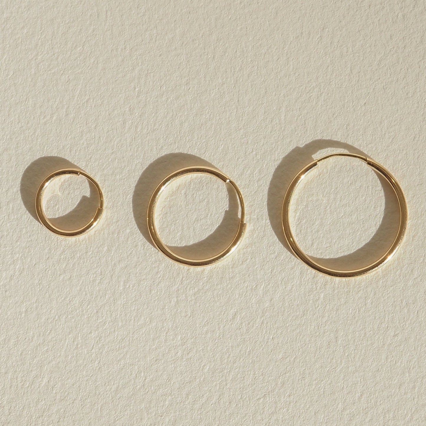 Four gold bangles of varying sizes displayed on a light textured surface, casting soft shadows, showcasing minimalist luxury jewellery design. [text overlay: 10mm (left), 15mm (middle) and 20mm (right)] [text colour: #000000]
