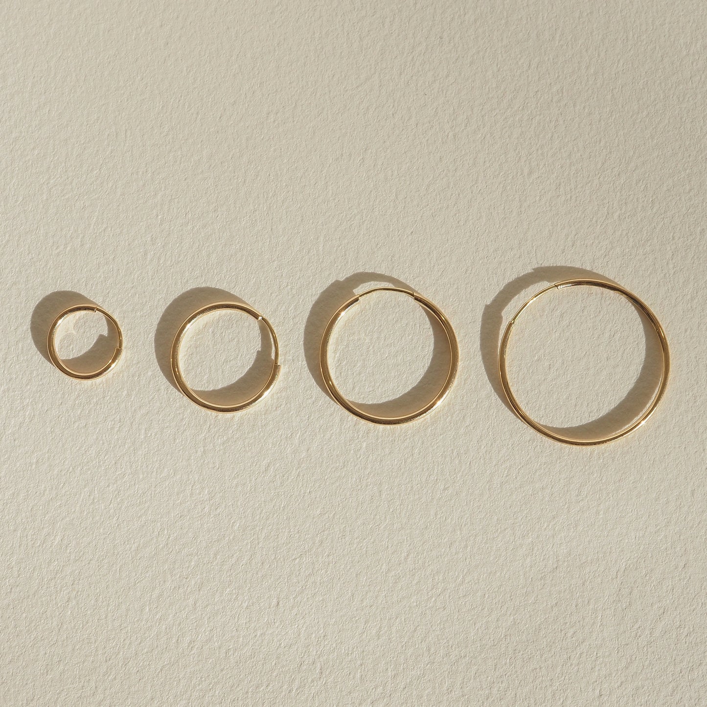 Four gold rings of varying sizes displayed on a textured beige surface, casting soft shadows. Elegant minimalist jewellery design.
