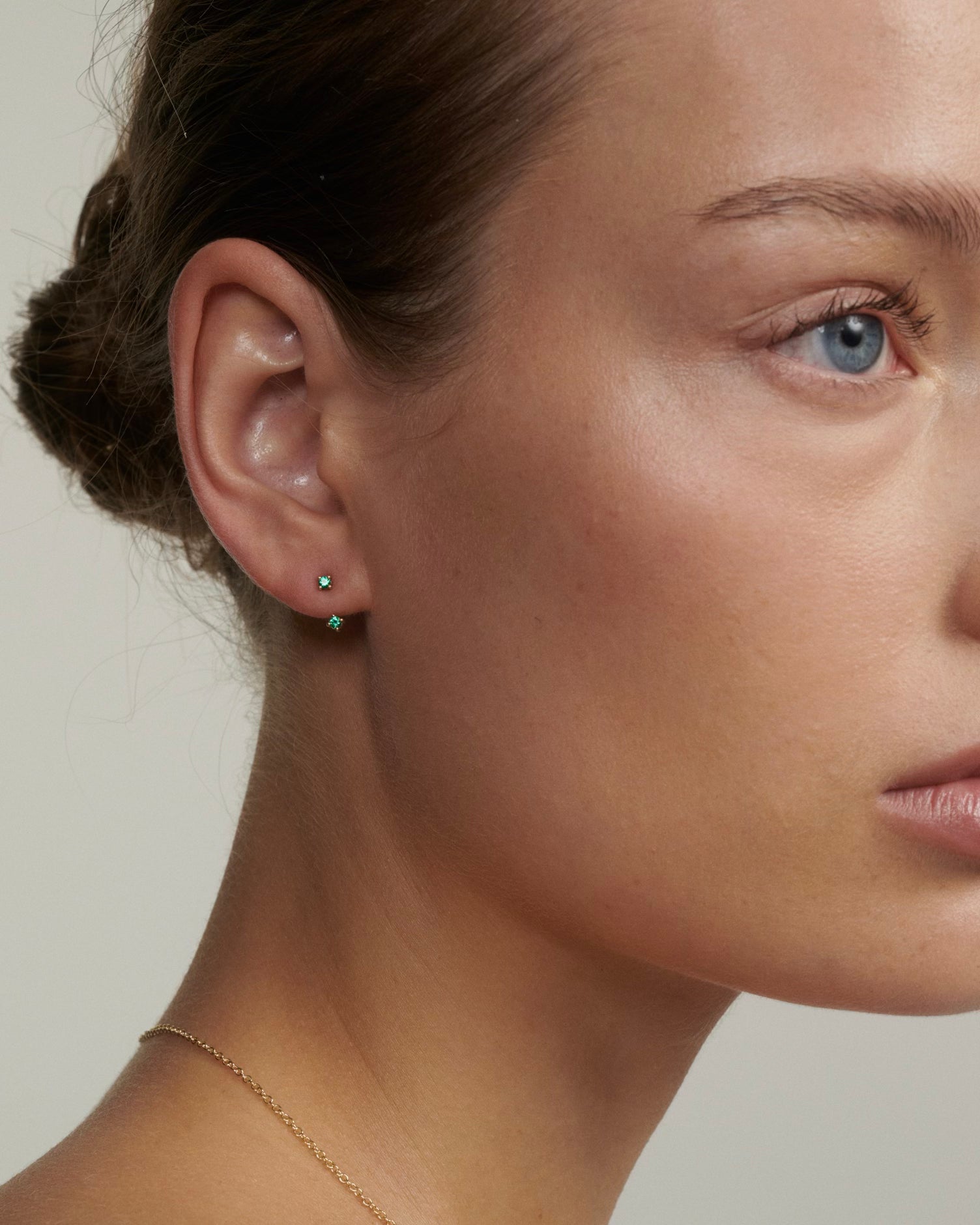 Close-up of a person wearing a green earring with a neutral background [text overlay: Andrea wears the Tiny Birthstone Jackets and Studs]