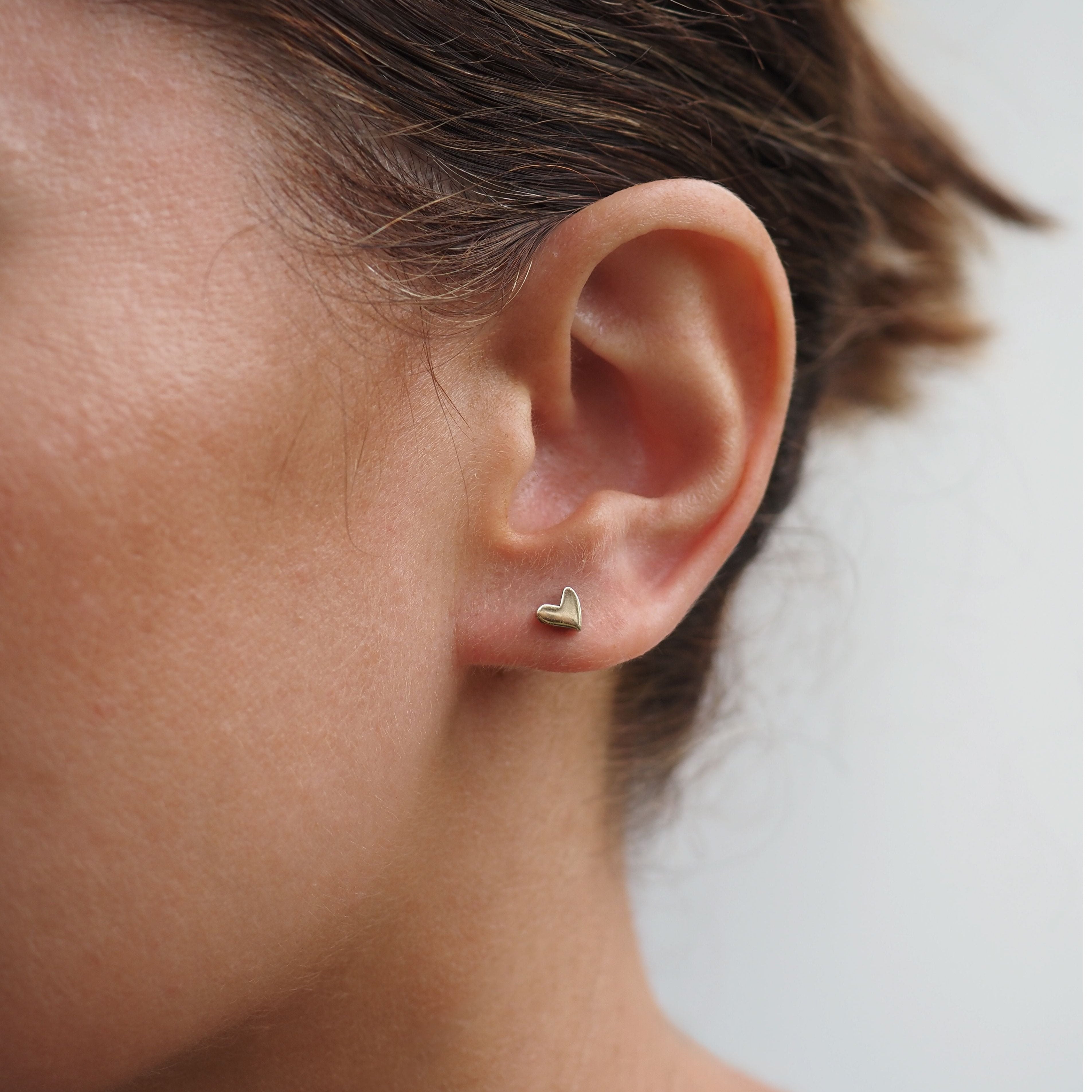 Close-up of a woman's ear wearing a small gold heart-shaped stud earring. Elegant and minimalist jewellery style.