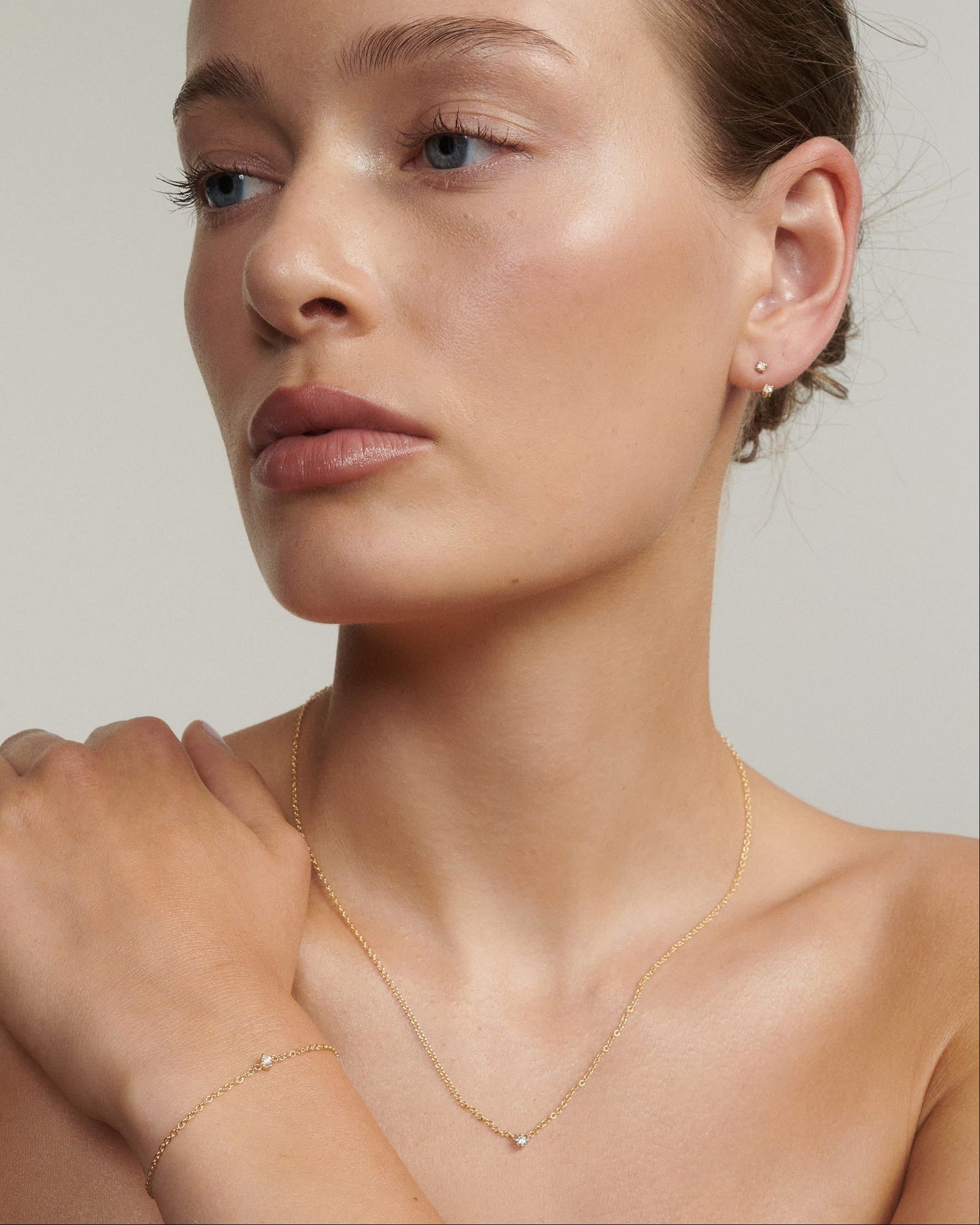 Woman wearing a delicate gold tiny birthstone necklace, tiny birthstone bracelet and tiny birthstone studs and jackets against a plain background