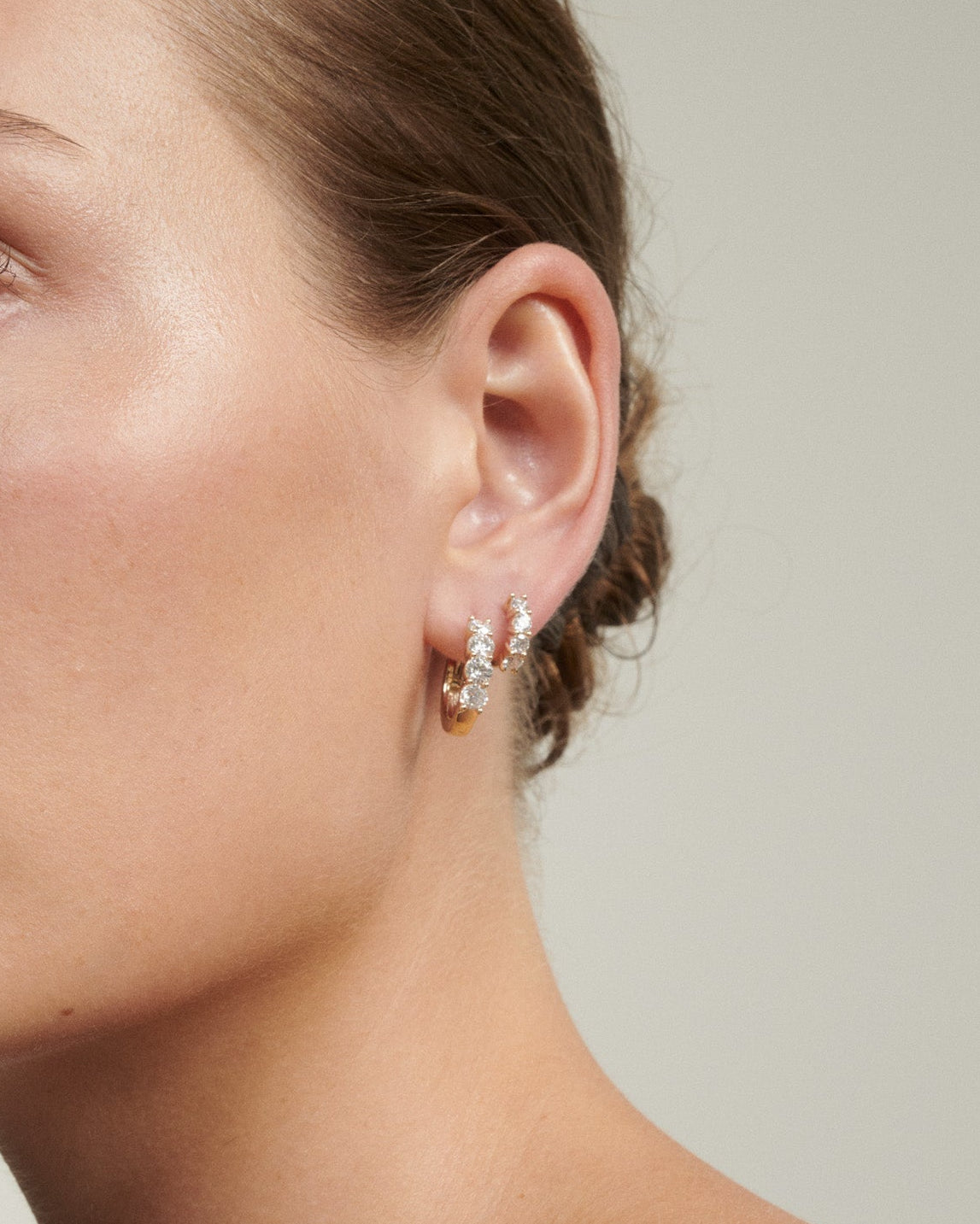 Close-up of a woman wearing a diamond earring against a neutral background [text overlay: Bold & Signature Diamond Huggies]