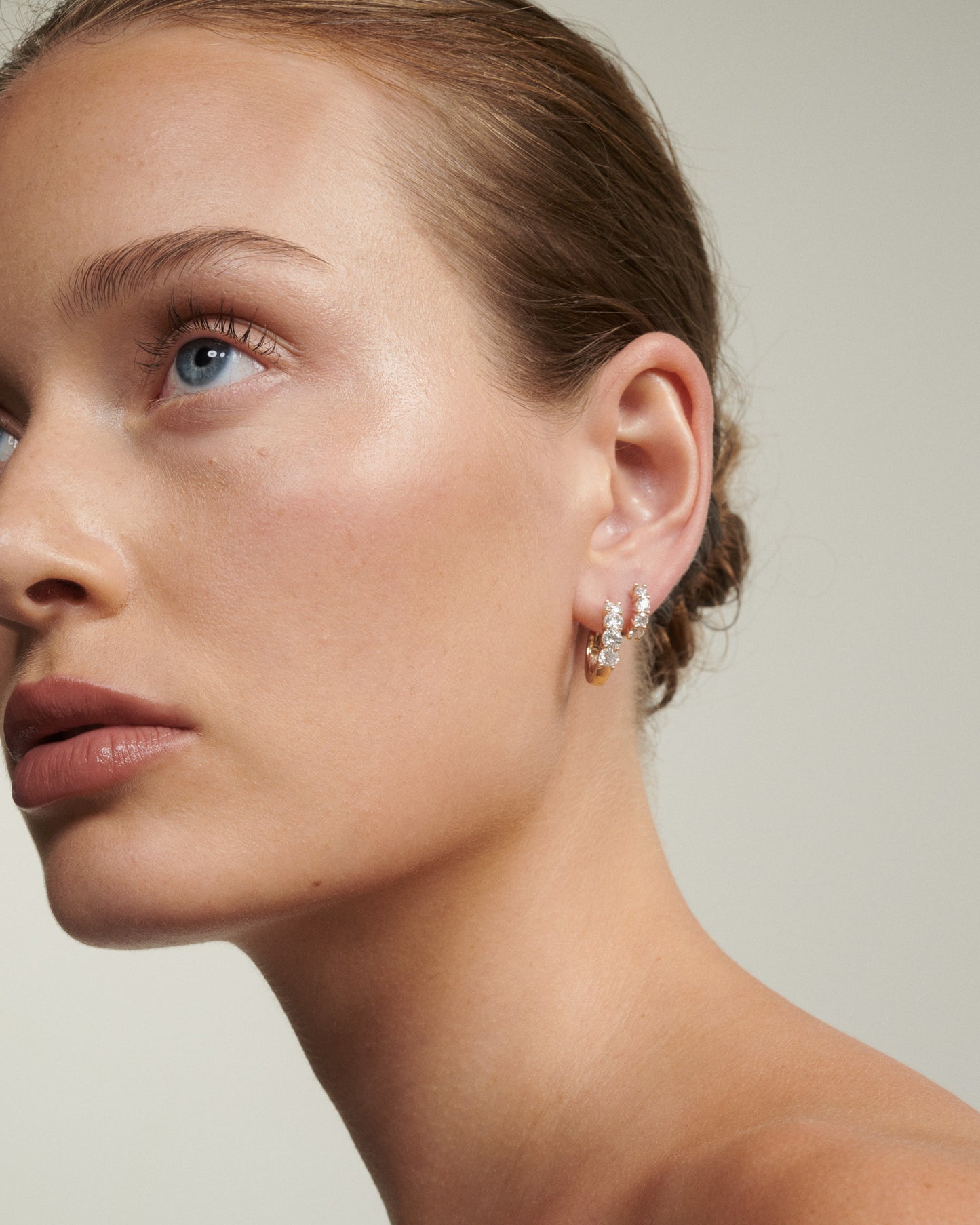 Close-up of a woman wearing a diamond earring against a neutral background [text overlay: Bold & Signature Diamond Huggies]
