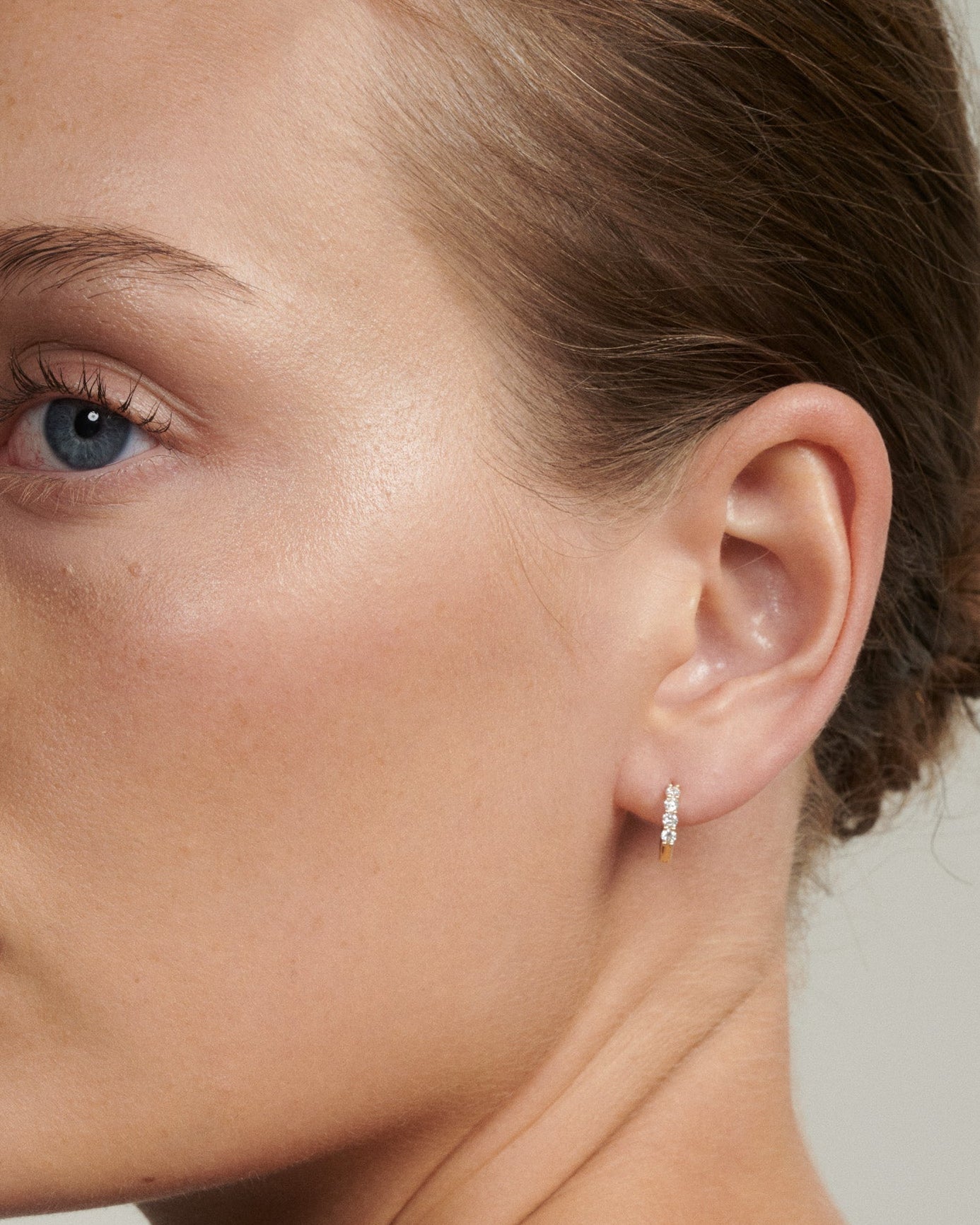 Close-up of a woman's ear wearing a diamond earring against a neutral background [text overlay: Tiny Diamond Huggies]