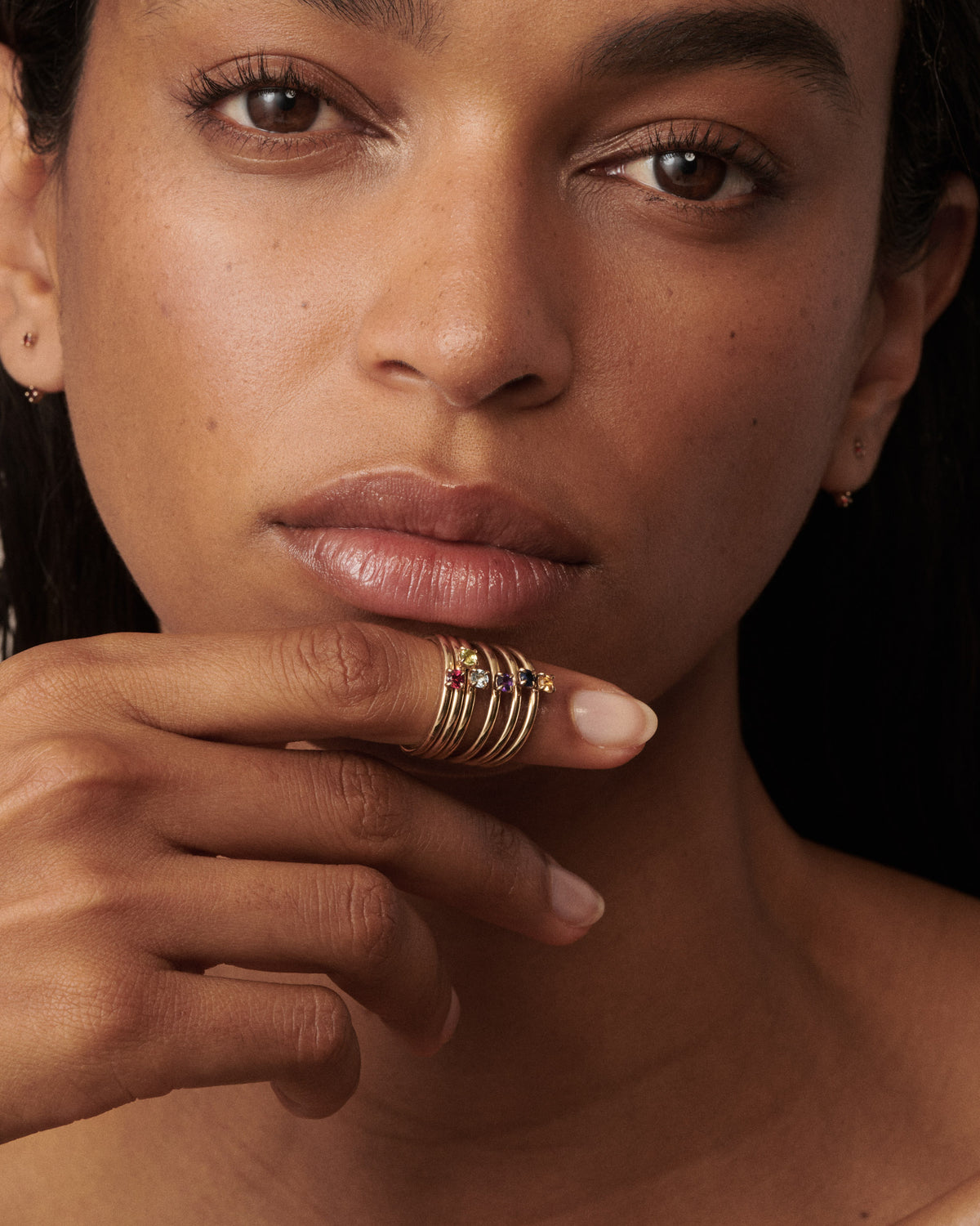 Woman wears a stack of 6 birthstone rings on her finger that is placing on her lips