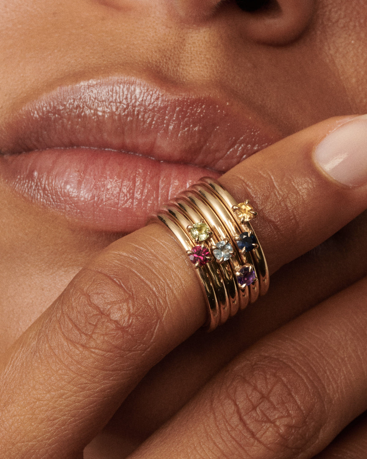 Woman wears a stack of 6 birthstone rings on her finger that is placing on her lips