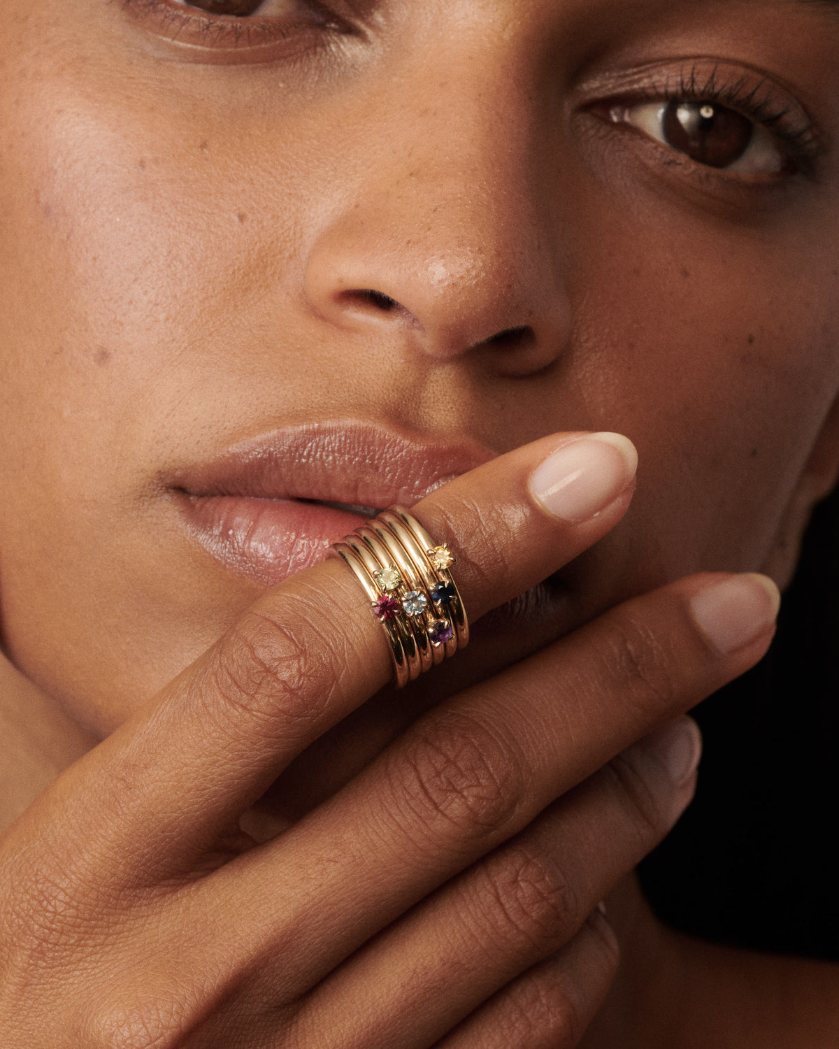 Woman wears a stack of 6 birthstone rings on her finger that is placing on her lips