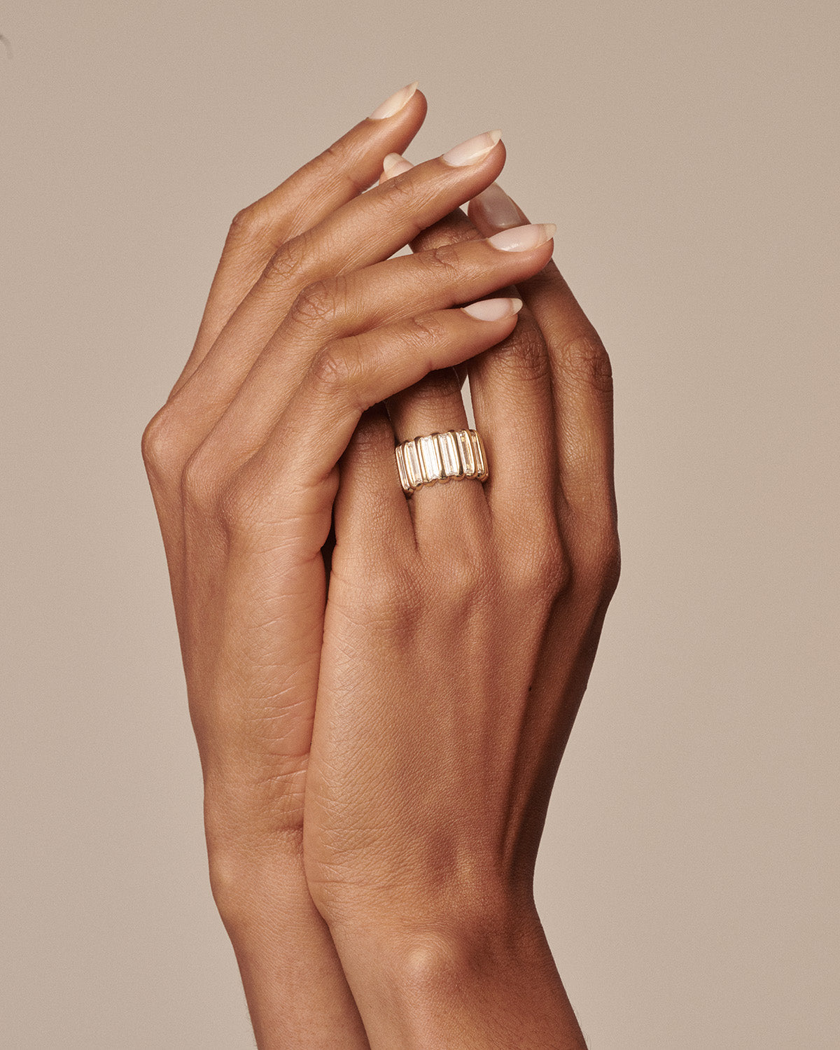 A close-up of a person's hands with manicured nails, wearing a wide gold ring with a textured ribbed design, set against a solid warm beige background.