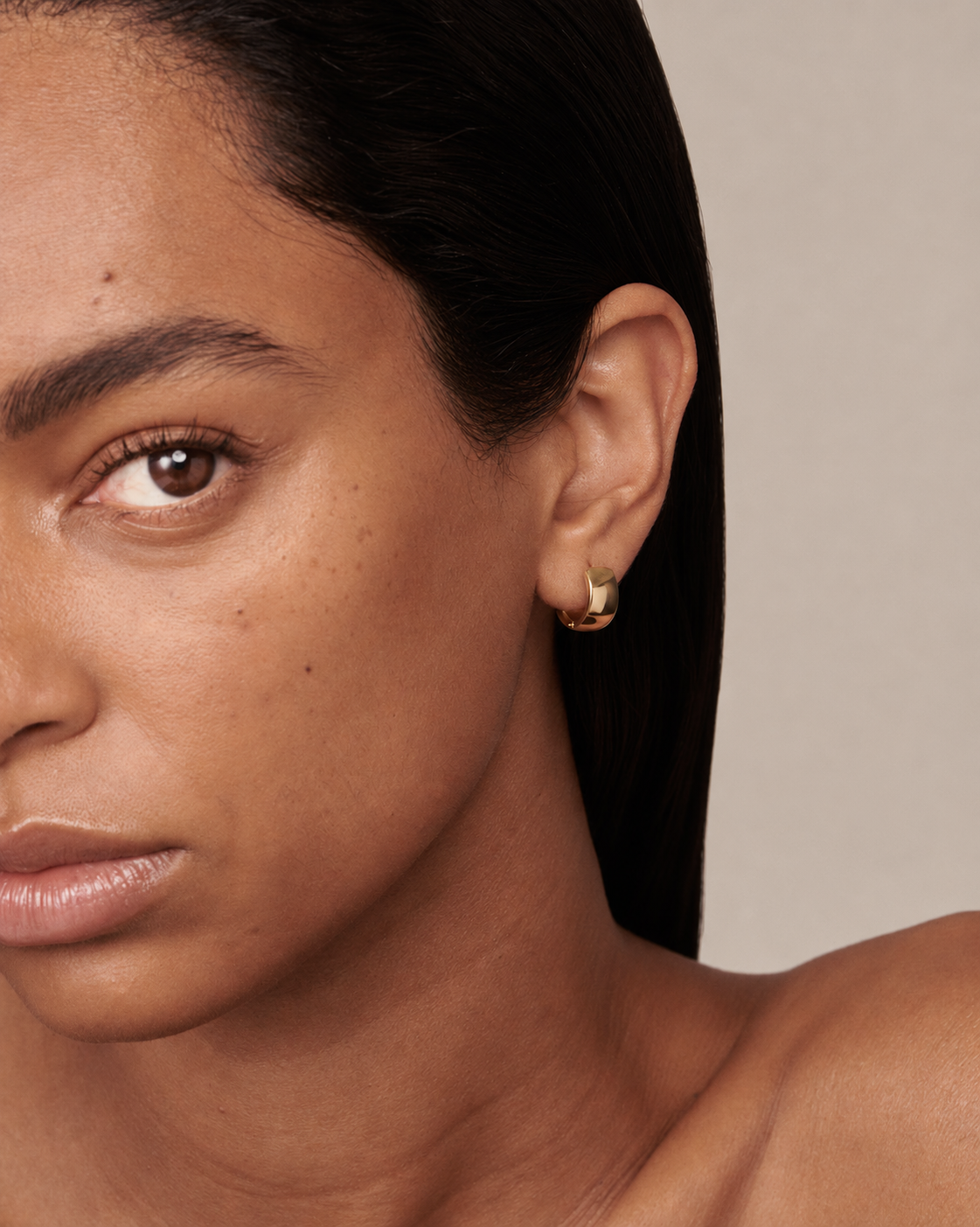 Close-up of a model wearing a small gold crescent moon hoop earring against a neutral beige background.